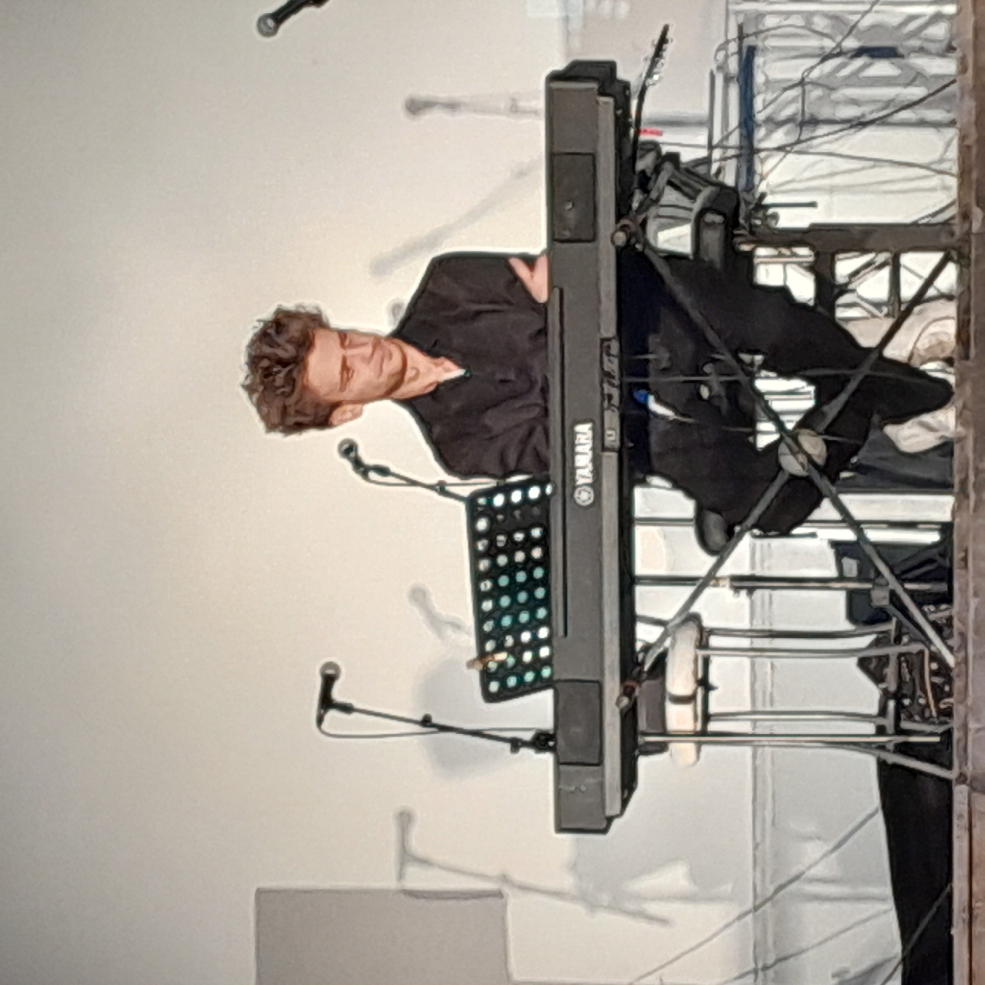 This image depicts a person playing a keyboard on a stage. The person, with curly brown hair and a light complexion, is wearing a dark collared shirt. They are seated behind a black Yamaha keyboard, which has a music stand attached. The keyboard is angled towards the viewer, and the music stand holds what appears to be a lit-up display with multiple circular lights.
The setting is indoors, likely a performance venue. The background is a light-colored wall. There are several microphones visible on stands around the stage. To the right of the keyboard, there is a metal structure, possibly part of a stage rig or scaffolding, with wires running across it. The lighting appears to be artificial, suggesting an indoor event.
The image is rotated 90 degrees clockwise, so the scene is presented sideways. The activity is clearly a musical performance. No specific time of day or weather conditions can be determined from the image. The focus is on the musician and their instrument, suggesting a portrait or candid shot of a performance. Visible text includes "YAMAHA" printed on the keyboard.
