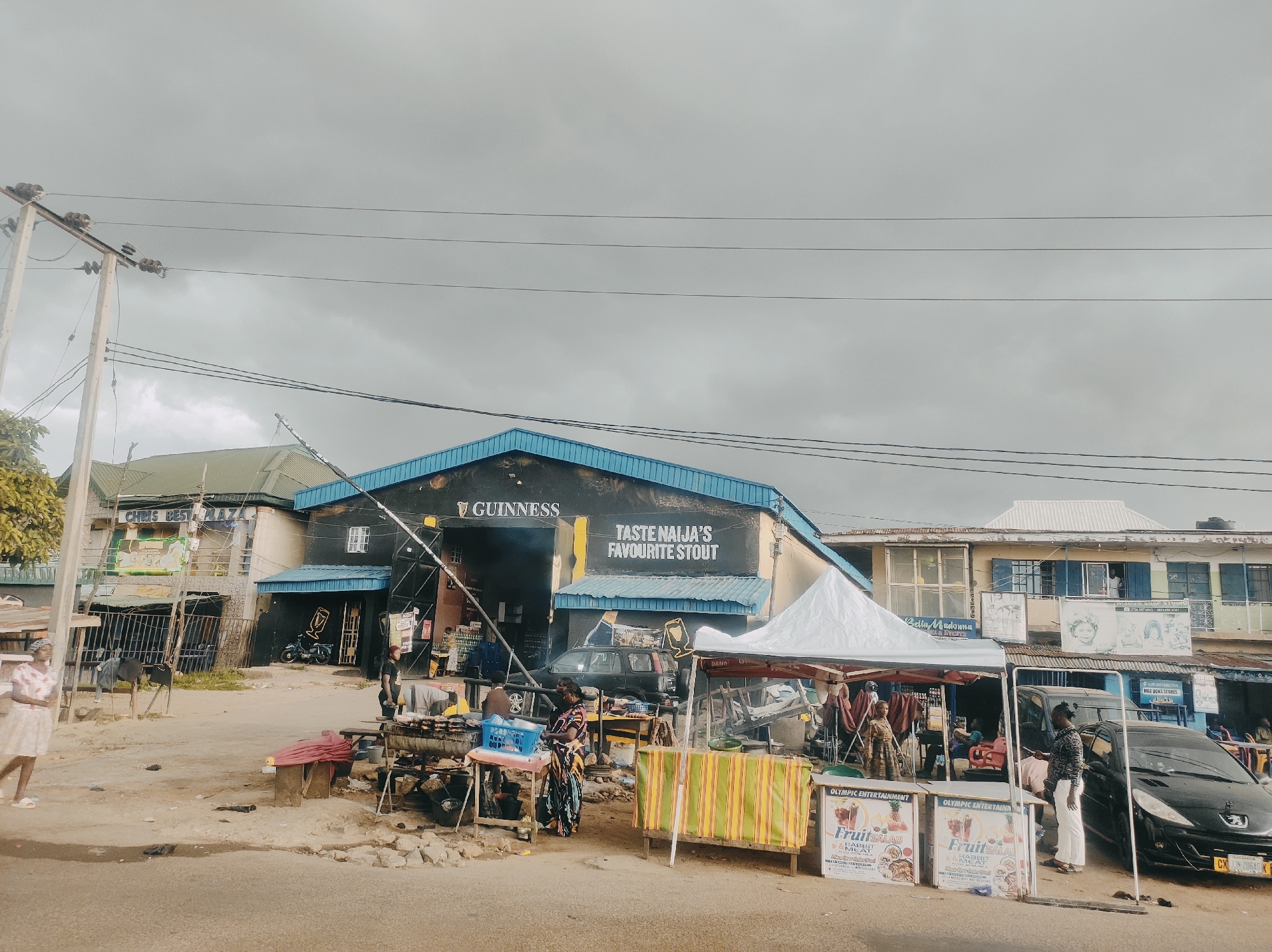 The image captures a street scene in Jos, Nigeria, under an overcast sky. A Guinness-branded building with the text "TASTE NAIJA'S FAVOURITE STOUT" prominently displayed is centrally located. Various commercial activities take place in front of the building, including food stalls under a white canopy, selling "Fruit Salad & Rabbit Meat." Several individuals are present, including vendors and customers, some engaged in transactions. A black car is parked beside the fruit stall, partially obstructing the view of the building behind. The environment appears to be a busy marketplace with other buildings visible in the background. A power pole with multiple wires stretches across the top of the image.
