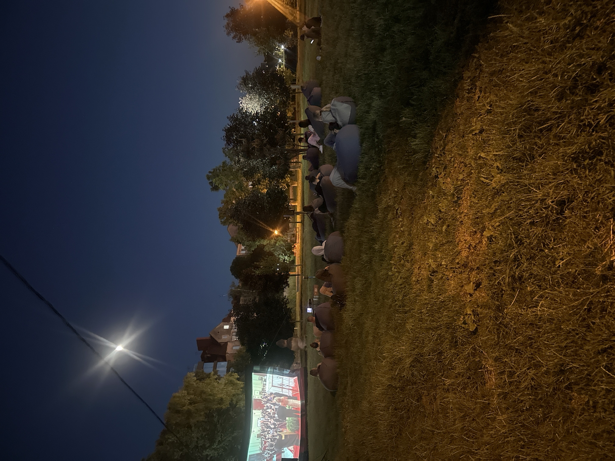 An outdoor movie screening is taking place at night on a grassy field in the city of Ternopil, Ukraine. A large projection screen displays a formal event, possibly a ceremony or a concert, with a seated audience visible on the screen. In the foreground, a group of people are seated on beanbag chairs on the grass, facing the screen. The sky is dark blue, with a bright moon visible in the upper left corner, casting a glow and appearing somewhat starburst-like due to the camera's aperture. Trees and buildings are visible in the background, with some streetlights providing illumination. The scene depicts a relaxed, communal gathering enjoying a film outdoors.