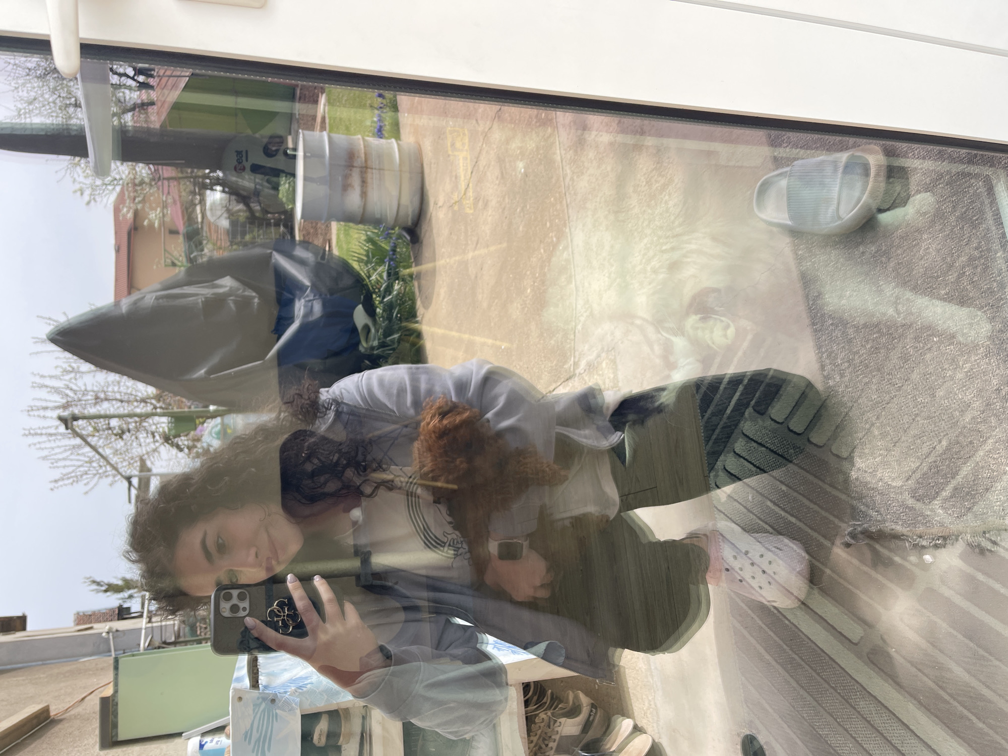 A reflection in a window shows a woman taking a selfie with her phone while holding a brown poodle. The background shows a backyard with a covered grill, a water barrel, and a building. A single sandal sits on the patio in the background, which is reflecting through the window. The location appears to be in a residential area in Sofia, Bulgaria.