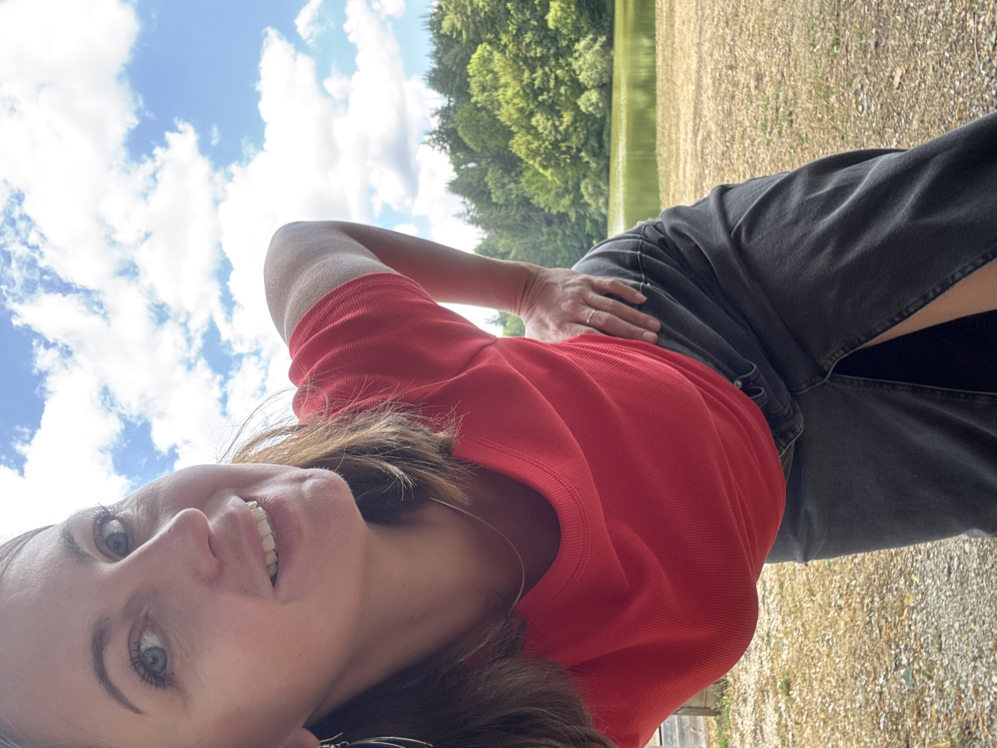 A woman is captured in a selfie, leaning towards the camera, with her head tilted and a smile on her face. She has brown hair and is wearing a red top and a dark skirt. Her right hand is resting on her hip, and a ring is visible on her finger. In the background, a body of water is surrounded by lush green trees under a partly cloudy blue sky. The ground in the foreground appears to be covered with gravel and dry grass. The scene suggests an outdoor setting, possibly a park or a lake in a rural area. The provided location, Coteaux du Lizon, France, is a municipality in the Jura department, known for its natural landscapes which would be consistent with the background of the image.
