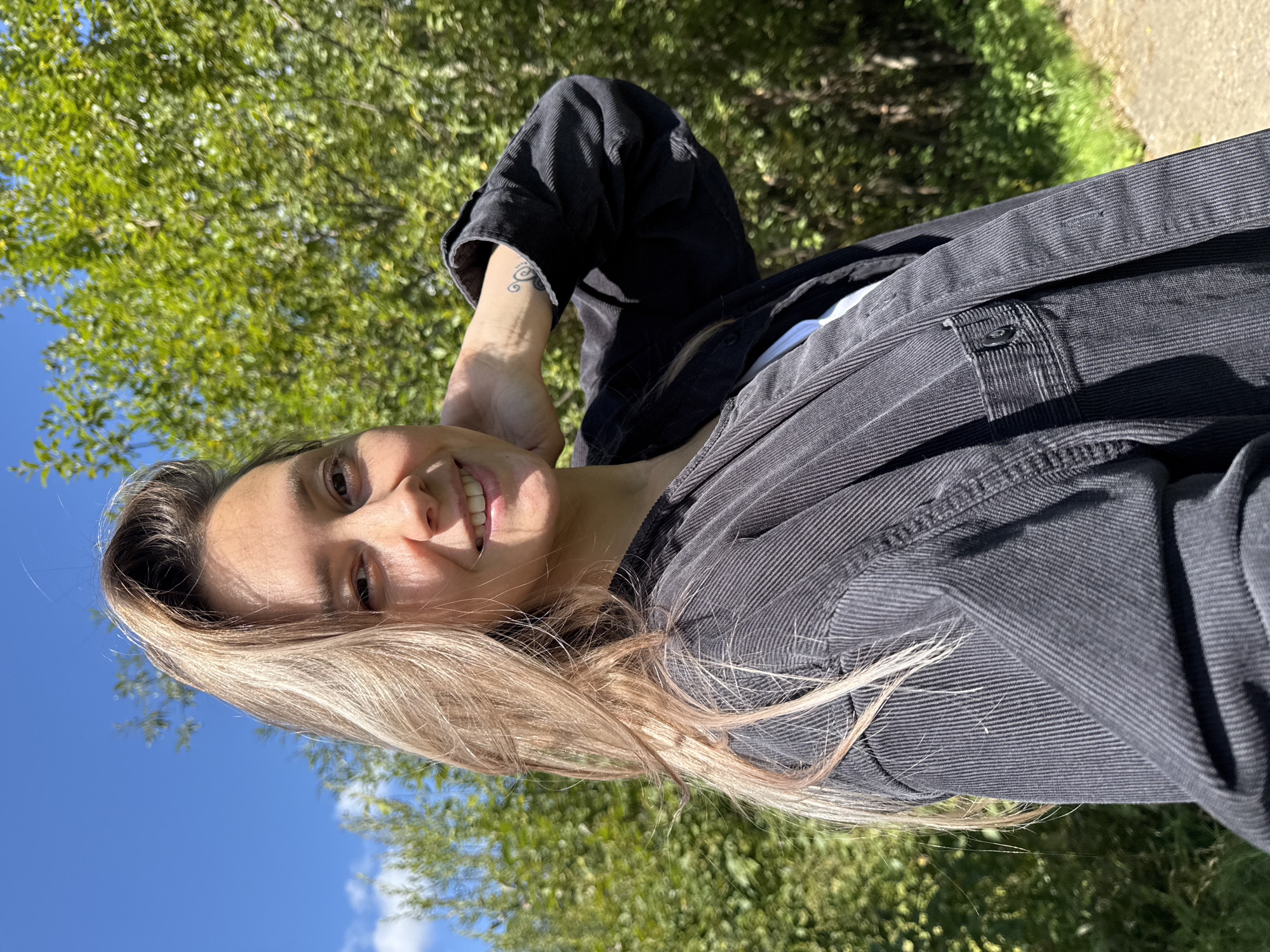 A young woman with blonde hair and a tattoo on her left arm is standing outdoors in front of a blurred background of green trees and a blue sky. She is wearing a dark corduroy jacket over a white t-shirt. She is smiling and looking towards the camera, with her right hand behind her neck. The lighting suggests it is daytime. The location is Gornozavodsk, Russia.
