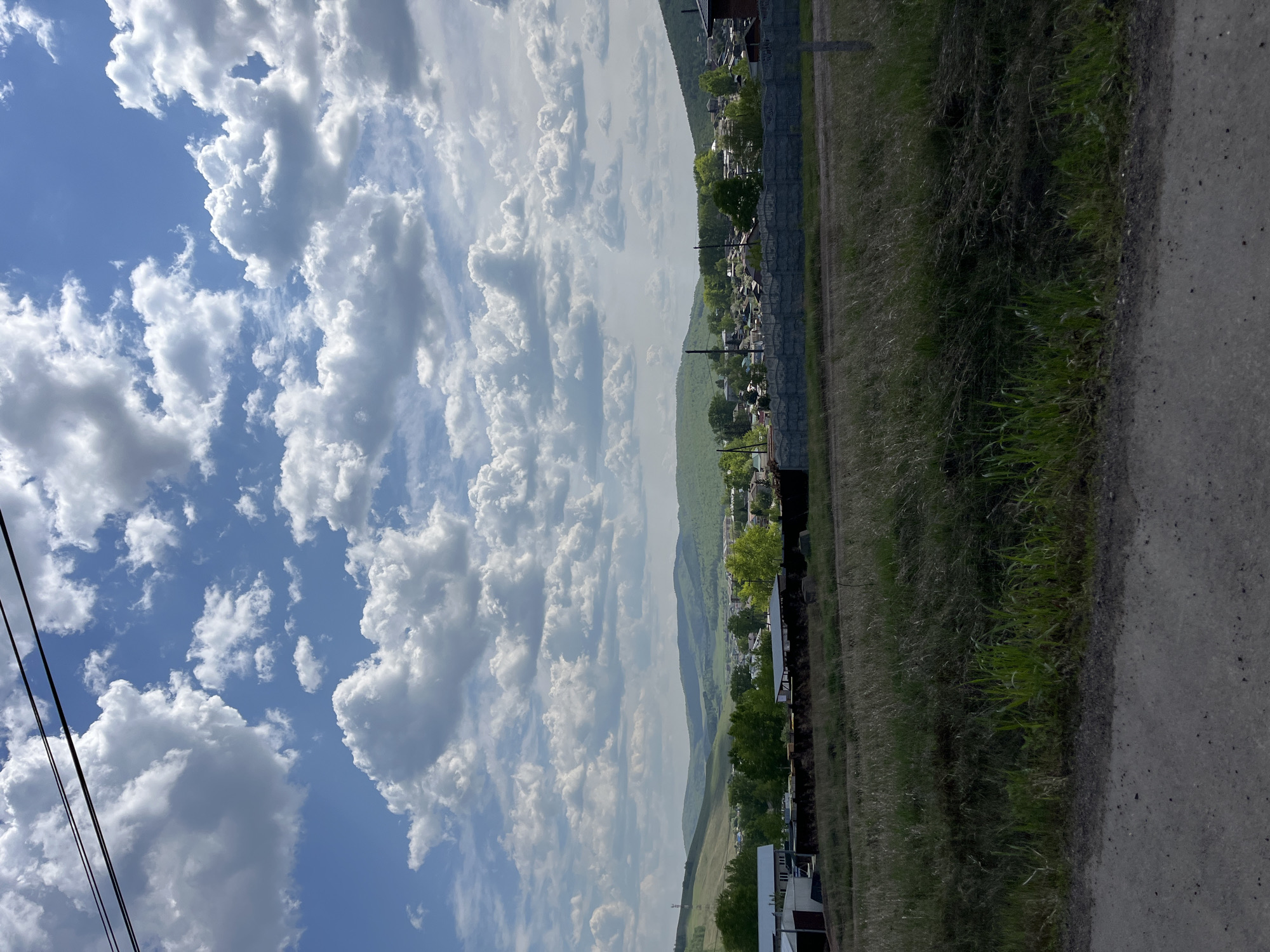 The image presents a landscape oriented vertically. Dominating the left portion is a sky filled with cumulus clouds set against a blue expanse. A mountainous terrain is visible, partially obscured by the clouds. The foreground features a dirt road bordering a field of grass. A fence runs parallel to the road, with glimpses of buildings interspersed with trees beyond the fence. The scene is located in the village of Зыково, Russia.