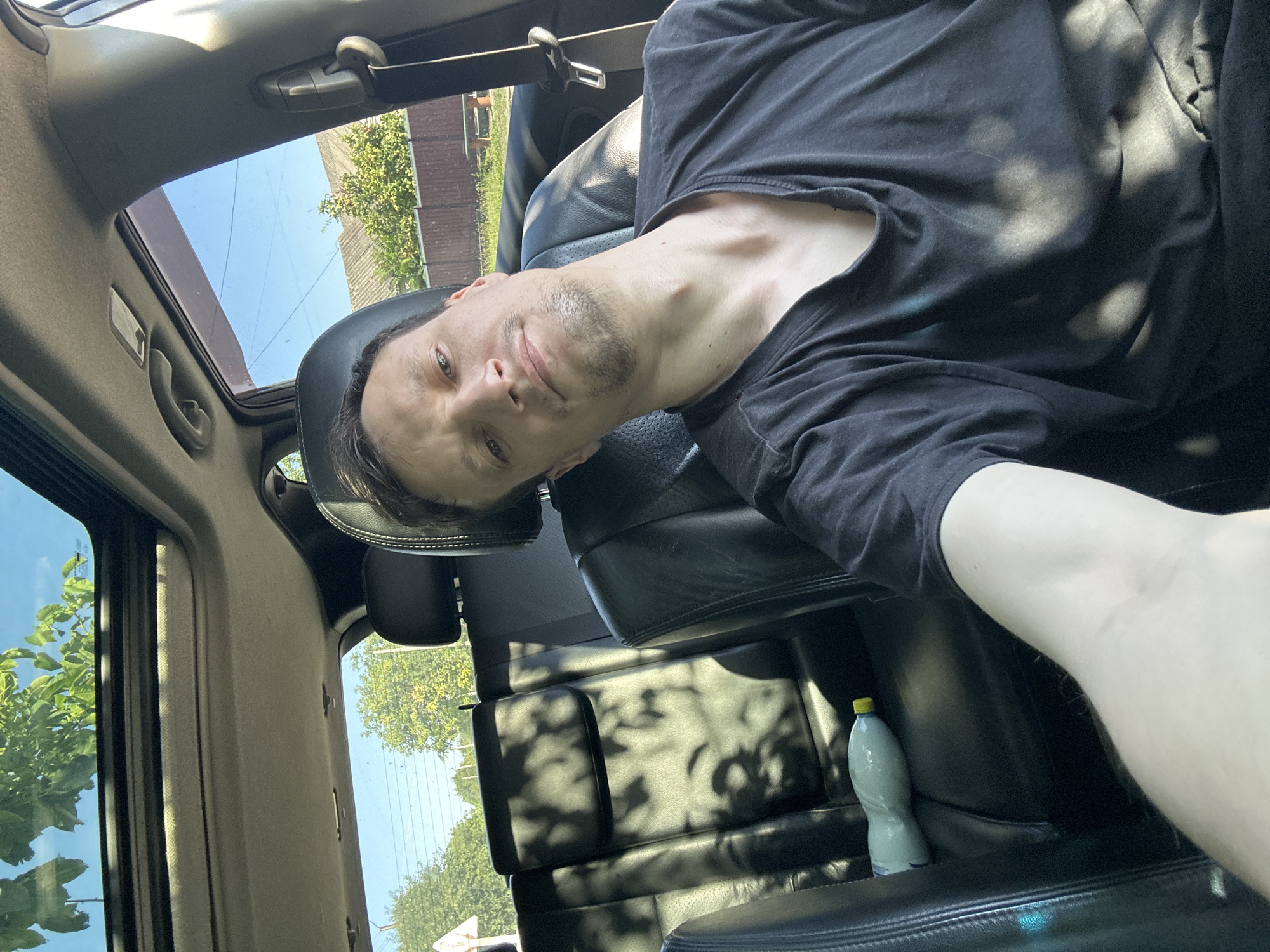 A man is taking a selfie in the passenger seat of a car, with the sunroof open. The car appears to be parked on a street in Shatava, Ukraine, as evidenced by the rural surroundings visible through the windows, including houses, fences, and trees. The man is wearing a black t-shirt and looking towards the camera with a neutral expression. A partially full plastic bottle with a yellow cap is visible on the back seat. Sunlight casts dappled shadows on the car's interior and the man's clothing.