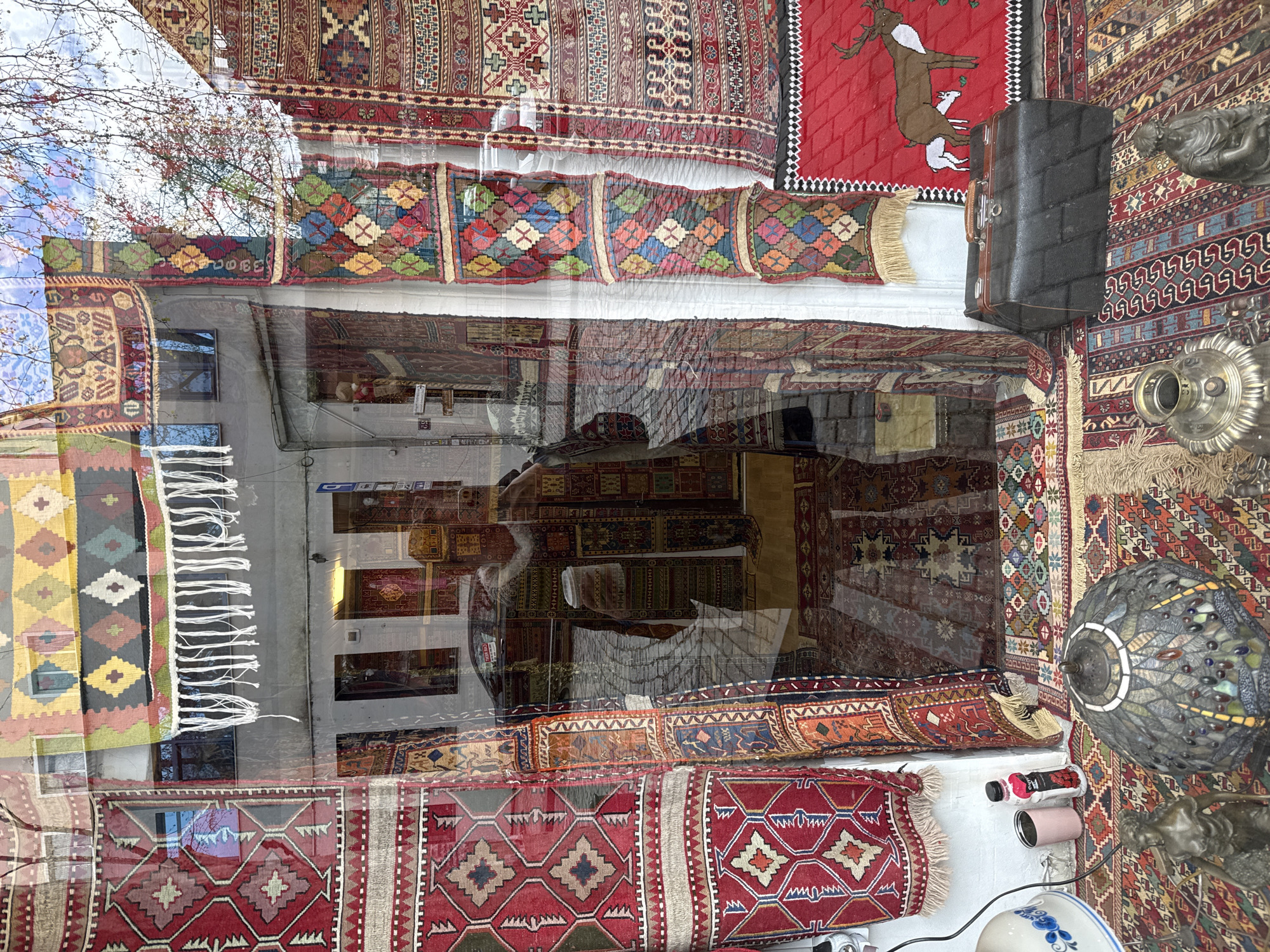 The image captures a storefront display of traditional rugs in Tbilisi, Georgia. A variety of colorful, patterned carpets are hung along the top of the store window and laid out inside. The reflections on the glass show an adjacent building with windows, hinting at an urban environment. Additional items, such as a vintage suitcase and decorative lamp, are also visible in the shop display. The scene suggests a shop specializing in handcrafted textiles and unique cultural items.