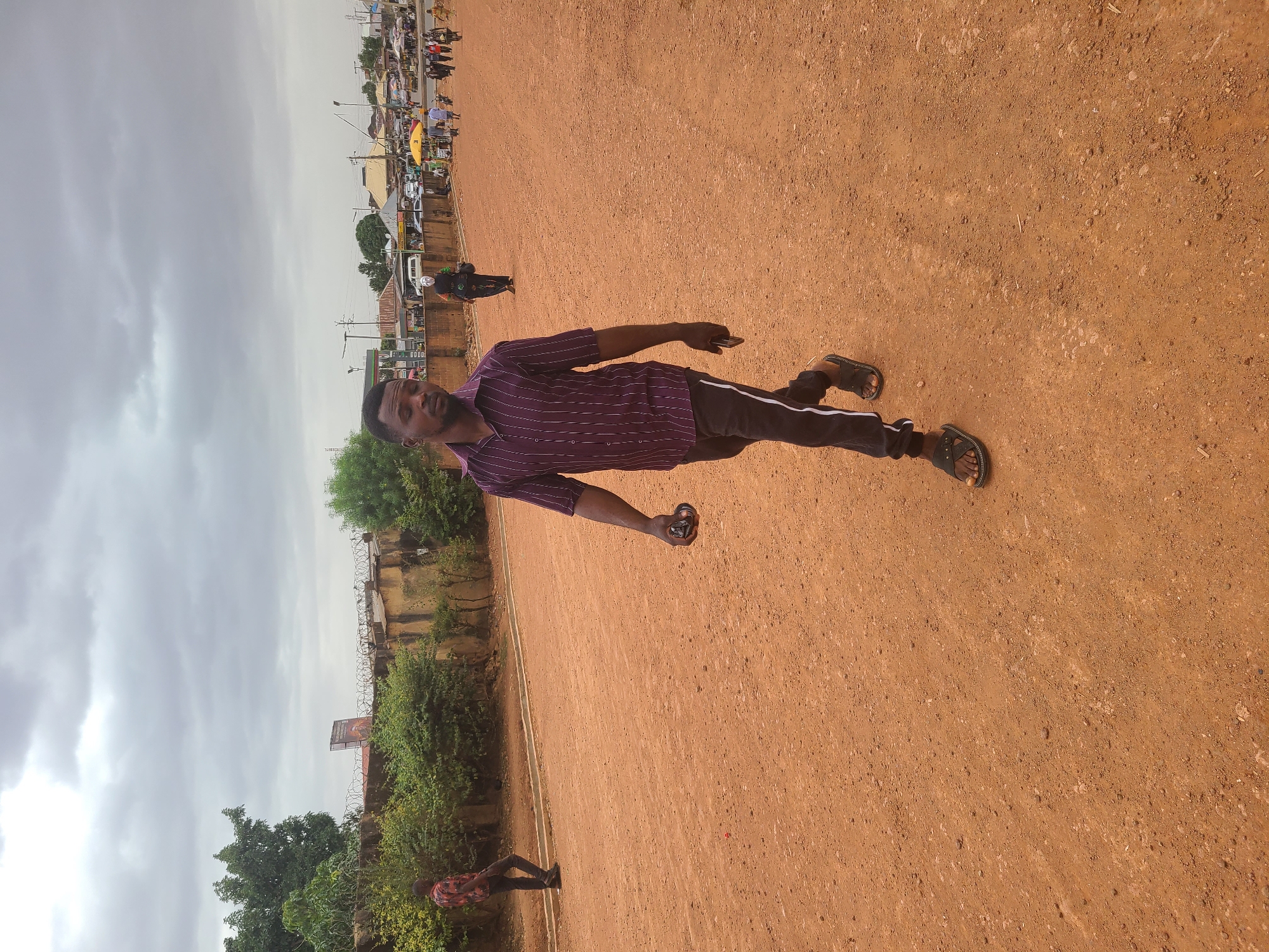 A man is standing on a dirt field in what appears to be Jos, Nigeria. He is wearing a purple striped shirt, black pants with a white stripe, and sandals. He is holding a small, round object in his right hand. In the background, there is a street with buildings and people. The sky is overcast. A fence is visible along the edge of the field with barbed wire.