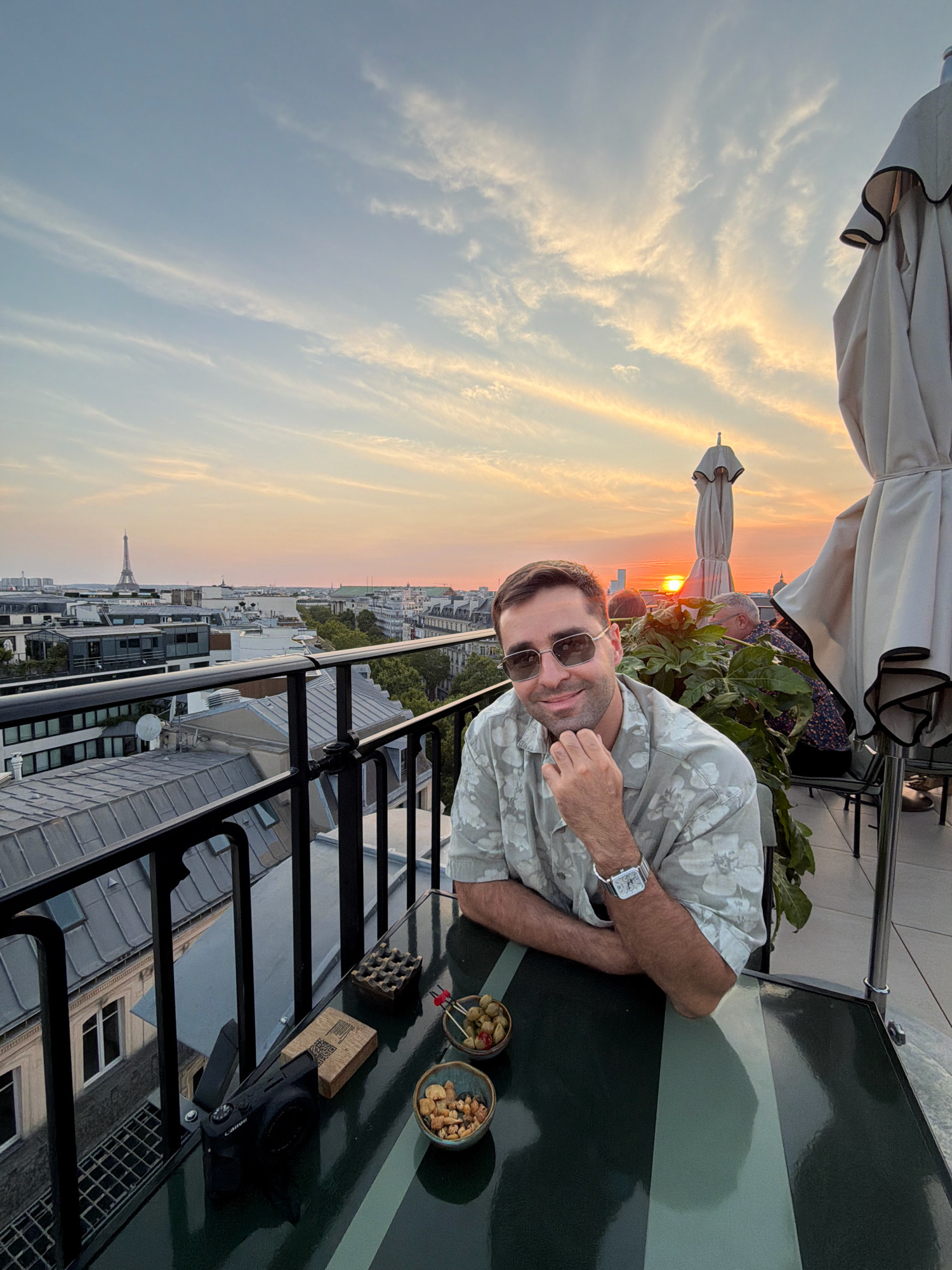 A man is seated at a table on a rooftop terrace overlooking Paris, France, during sunset. The Eiffel Tower is visible in the distance. The man is wearing sunglasses and a floral shirt, and he is resting his chin on his hand. On the table in front of him are a camera, an ashtray, and two bowls of snacks. Two outdoor umbrellas are also visible on the terrace. The sky is filled with soft clouds illuminated by the setting sun.