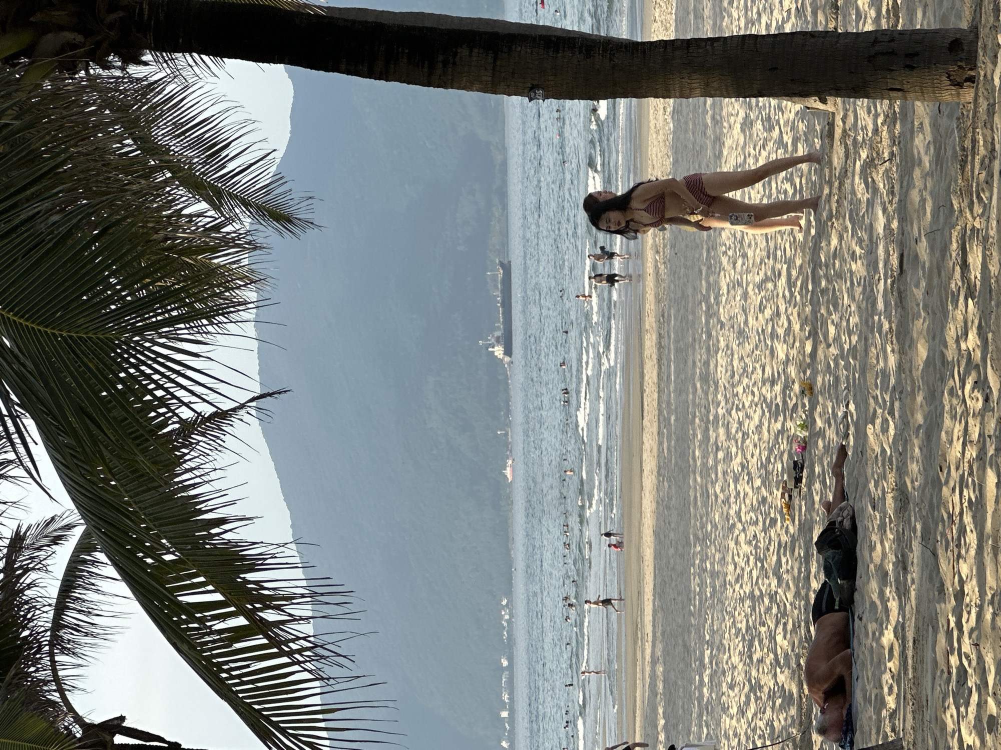 An aerial view of a beach in Da Nang, Vietnam. The beach is sandy and flat, with a few palm trees on the right side of the frame. In the foreground, a man is sleeping on the sand. In the middle ground, two young women are walking on the beach. In the background, there are several people swimming in the ocean and a cargo ship is visible on the water. The ocean is blue and calm, with small waves. In the distance, there are mountains. The sky is clear and overcast. The scene appears to be a typical summer day at the beach.