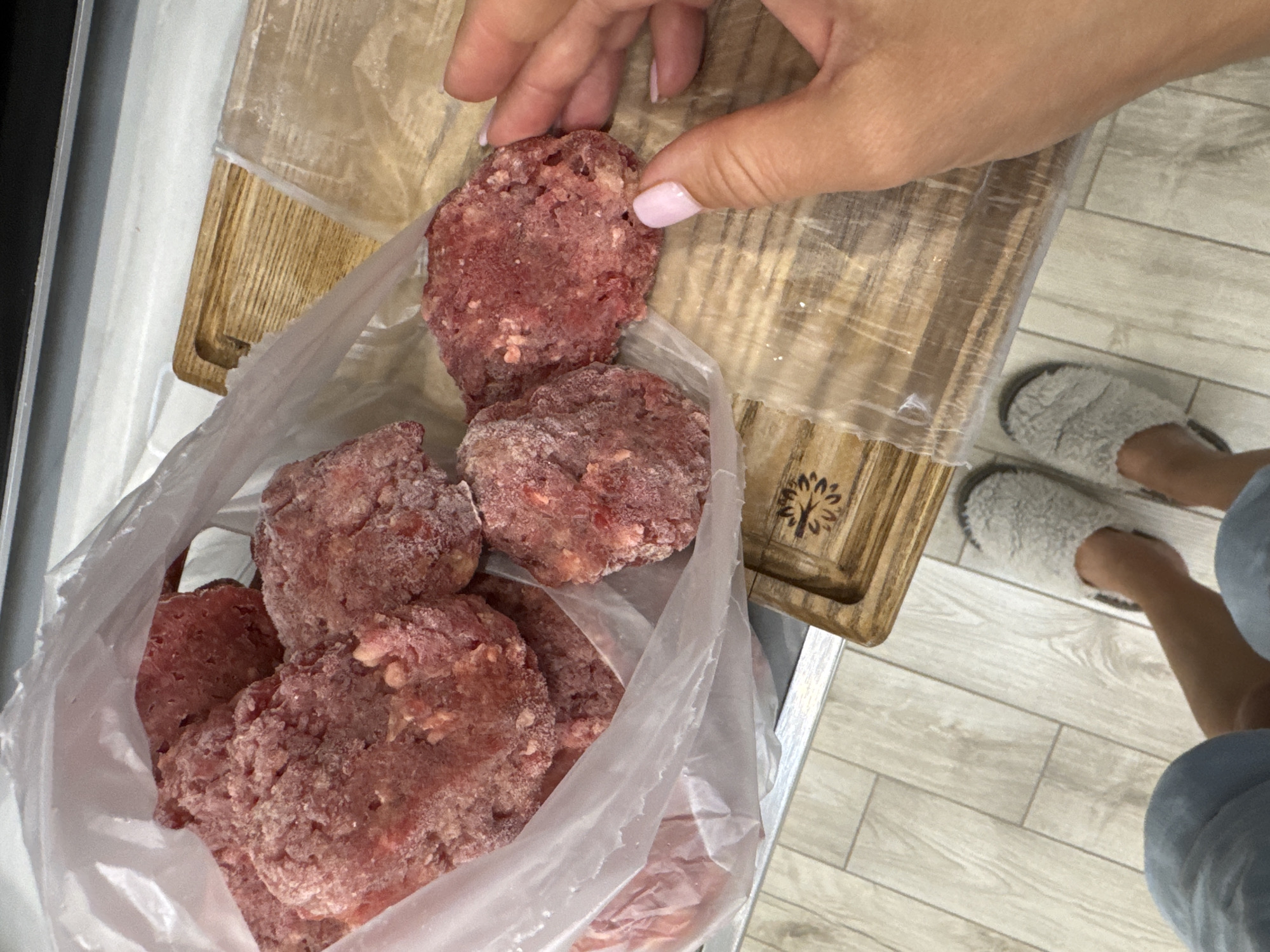 A person's hand with light pink nail polish is reaching for a frozen hamburger patty from a clear plastic bag filled with more frozen patties. The patties are raw ground meat, roughly formed into discs. They are sitting on a wooden cutting board, which has a transparent plastic sheet over it. In the background, a grey wooden floor is visible, and the person is wearing fluffy grey slippers. The scene appears to be indoors, likely a kitchen. No specific location in Sochi, Russia can be determined from the image.