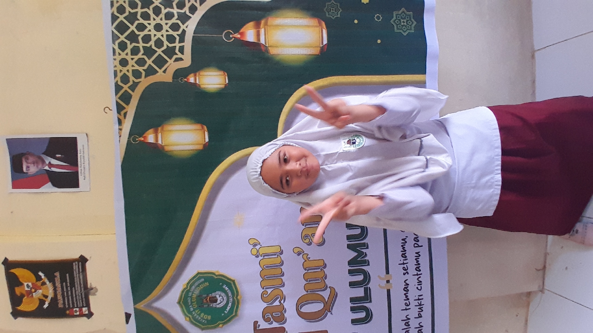 A young girl wearing a white headscarf and maroon and white uniform makes peace signs with both hands while posing in front of a banner in a room. The banner reads "Tasmi 'ul Qur'ai ULUMU" with the text below it in Indonesian. The banner contains a logo that includes the text "YAYASAN BAHADURUDDIN", "BDS IT" and "+ KHOKSEUMAWE" in a circular pattern. On the wall to the left, there are two pictures: one of an Indonesian official and another of the Indonesian coat of arms, Garuda Pancasila. The location is likely in Lhokseumawe, Indonesia, based on the writing on the banner.