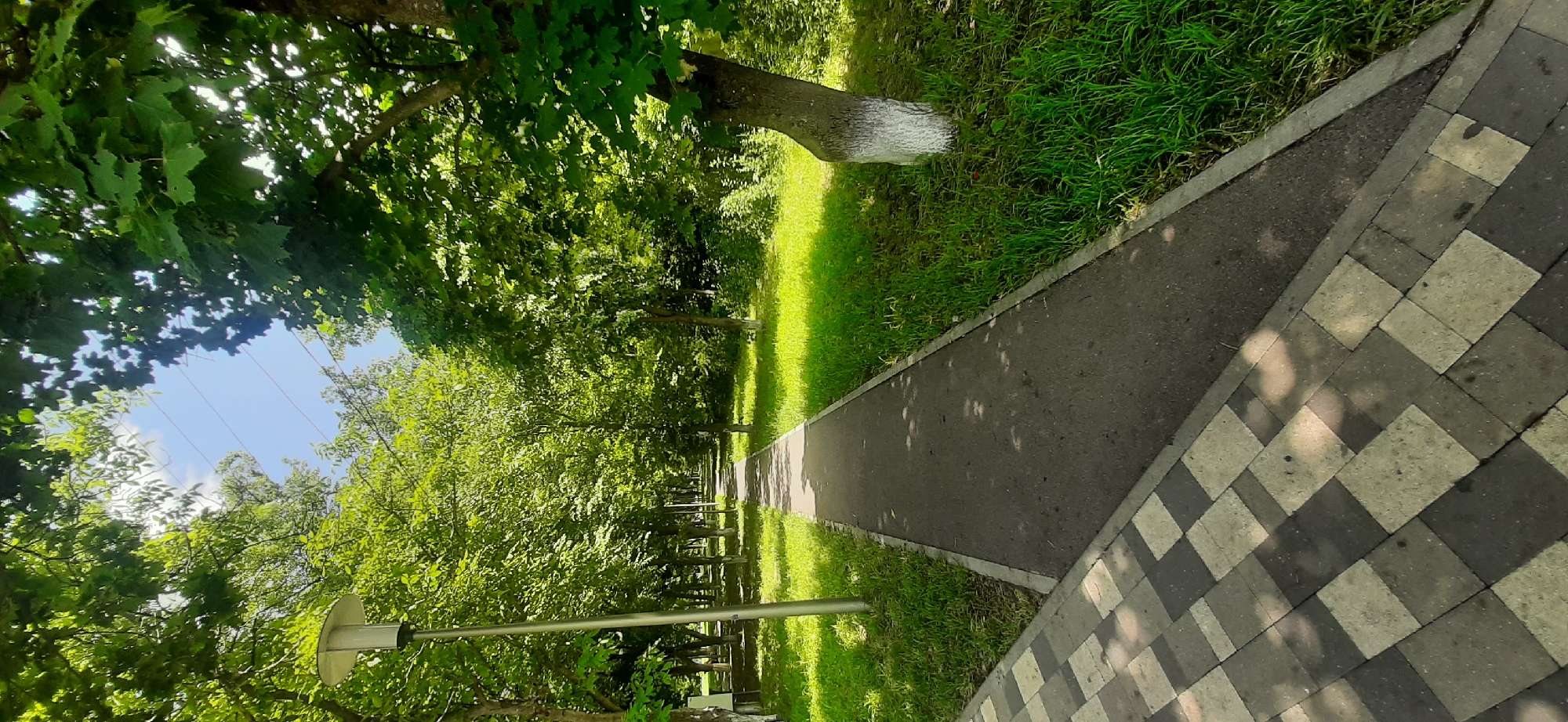 A paved pathway winds through a park lined with lush green trees and grass. The pathway is made of asphalt and bordered by a light grey concrete edge. To the left, a section of the pathway is paved with grey and beige square tiles laid in a checkered pattern. A modern street lamp stands tall on the left side of the asphalt path. The trees have dense green foliage, and sunlight filters through the leaves, casting dappled shadows on the ground. In the background, power lines are visible against a bright blue sky. The scene is one of a well-maintained public park in Yessentukskaya, Russia, on a sunny day.