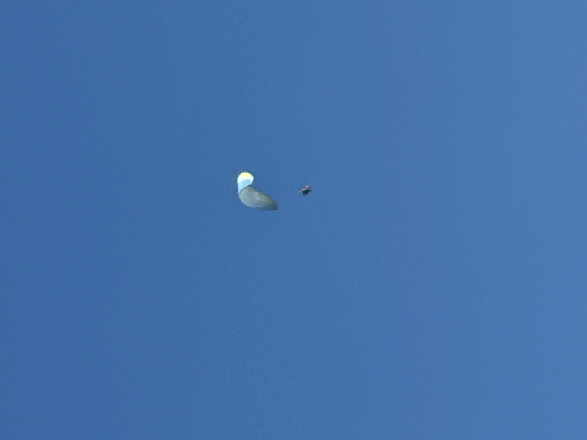 A single paraglider is visible in a clear blue sky, appearing as a small dark silhouette beneath a colorful paraglider. The paraglider itself has a curved wing, predominantly dark green or blue, with a bright yellow and white section on its leading edge. The paraglider is positioned in the upper right quadrant of the image, with the person underneath it slightly to the left and below the wing. The environment is a vast expanse of uninterrupted blue sky, with no discernible features of the city of Boreti, Montenegro visible. The lighting suggests it is daytime, with no shadows or specific light sources apparent within the frame.
