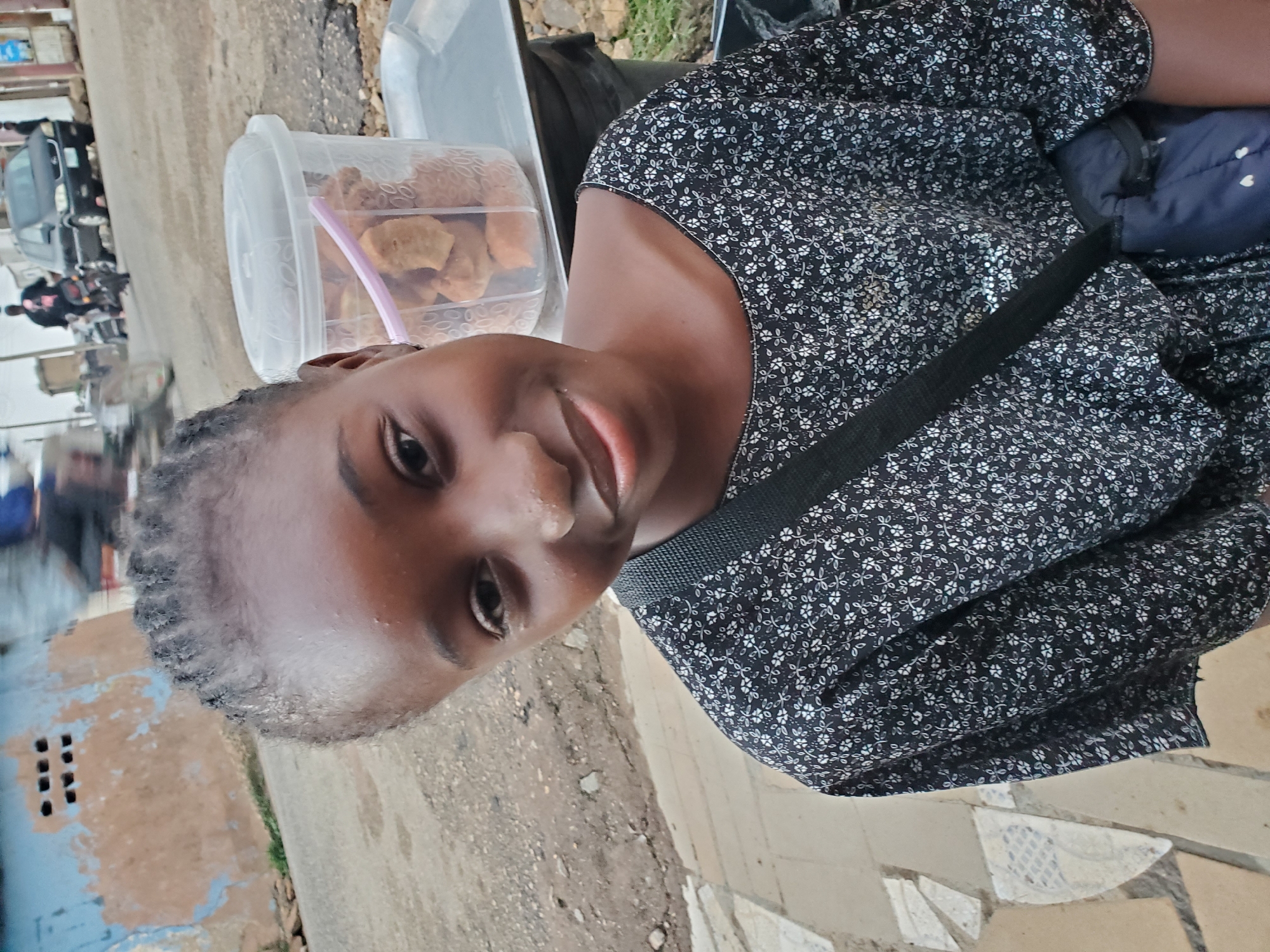 This is a vertical, eye-level, medium shot photograph of a young Black girl posing for the camera. She is wearing a dark-colored, short-sleeved dress with a white floral pattern. A dark strap, possibly from a bag, is visible across her chest. She has her hair braided into cornrows and appears to be looking directly at the camera with a neutral expression. Her skin is dark and smooth.
To her left, a clear plastic container is filled with what appear to be snacks, possibly fried dough or biscuits, with a pink straw sticking out. In the background, out of focus, there are hints of a street scene with vehicles and a building. The ground appears to be a mix of concrete and dirt. The overall lighting suggests it is daytime, with no clear indication of the weather. The setting appears to be an outdoor, possibly urban, environment. Given the context of Jos, Nigeria, it is likely a street or market scene. There is no visible text in the image.
