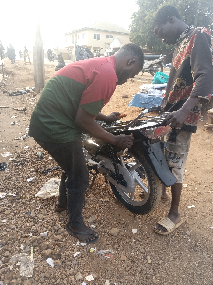 Two men are repairing a motorcycle on a dirt road littered with trash. One man wears a red and green shirt and jeans, working on the bike with a tool. The other man, wearing a patterned shirt and shorts, assists. The motorcycle is black and silver. A building is in the background. Other people and parked motorcycles are visible in the distance.