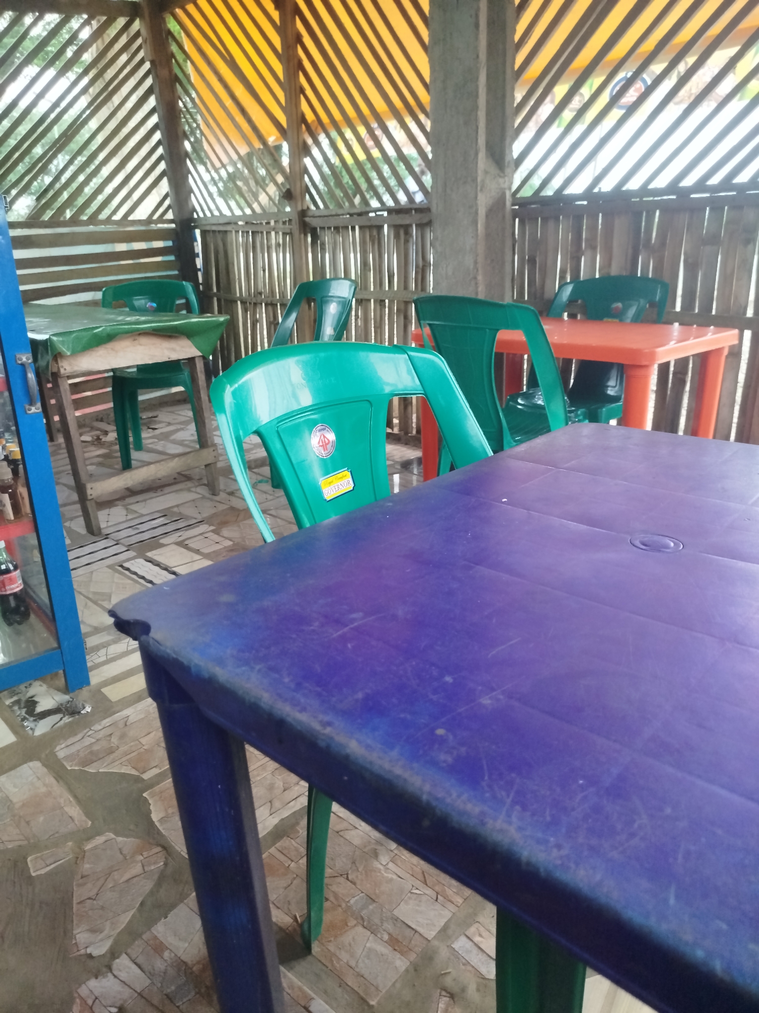 The image depicts an outdoor seating area with tables and chairs, likely a restaurant or roadside eatery, in Yenegoa, Nigeria. The seating area is covered by a slanted roof made of diagonal wooden slats and an orange tarp. The walls are constructed from vertical bamboo poles. In the foreground, a weathered purple plastic table is visible, with its blue legs extending downwards. To the left of this table, a green plastic chair is partially visible. In the middle ground, a green plastic chair with a white sticker that reads "GOVERNOR" is prominently displayed. Behind it, another green chair is tucked under an orange plastic table. Further back, a wooden table covered with a green plastic sheet is set with chairs. A blue display cabinet with bottles of drinks is on the far left. The floor is paved with irregular stone tiles. The overall scene suggests a casual, open-air dining establishment.