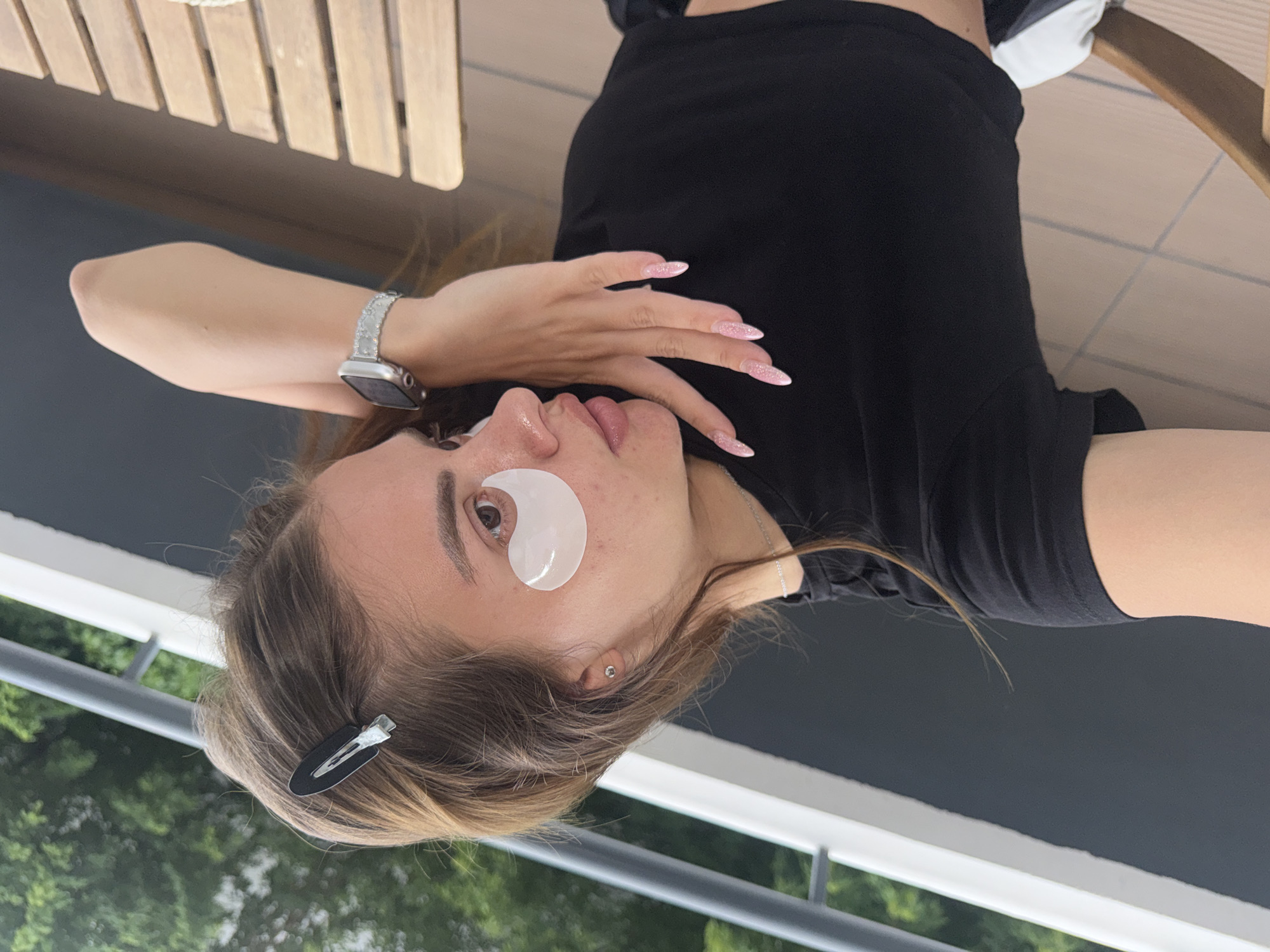 A young woman is depicted in a close-up, rotated selfie-style image, appearing to be reclining or lying down. She has light brown hair, which is partially secured with a black hair clip on the left side of her head. She is wearing a black short-sleeved t-shirt and a silver chain necklace. On her left wrist, she sports an Apple Watch with a decorative, possibly diamond-encrusted, silver band. Her long fingernails are painted with a pink glitter polish.
A white, crescent-shaped hydrogel patch is applied under her right eye, indicating a skincare routine. A small silver stud earring is visible in her right ear. Her lips are slightly parted, and her gaze is directed towards the camera with a relaxed expression.
The background features elements suggestive of an outdoor setting, likely a balcony or patio. Wooden slatted furniture, possibly a chair or table, is visible in the upper part of the frame. A dark grey or black surface, possibly a railing or floor, extends across the middle. Below this, blurred green foliage indicates the presence of trees or bushes. The lighting is soft and natural, suggesting daytime. There is no visible text in the image.