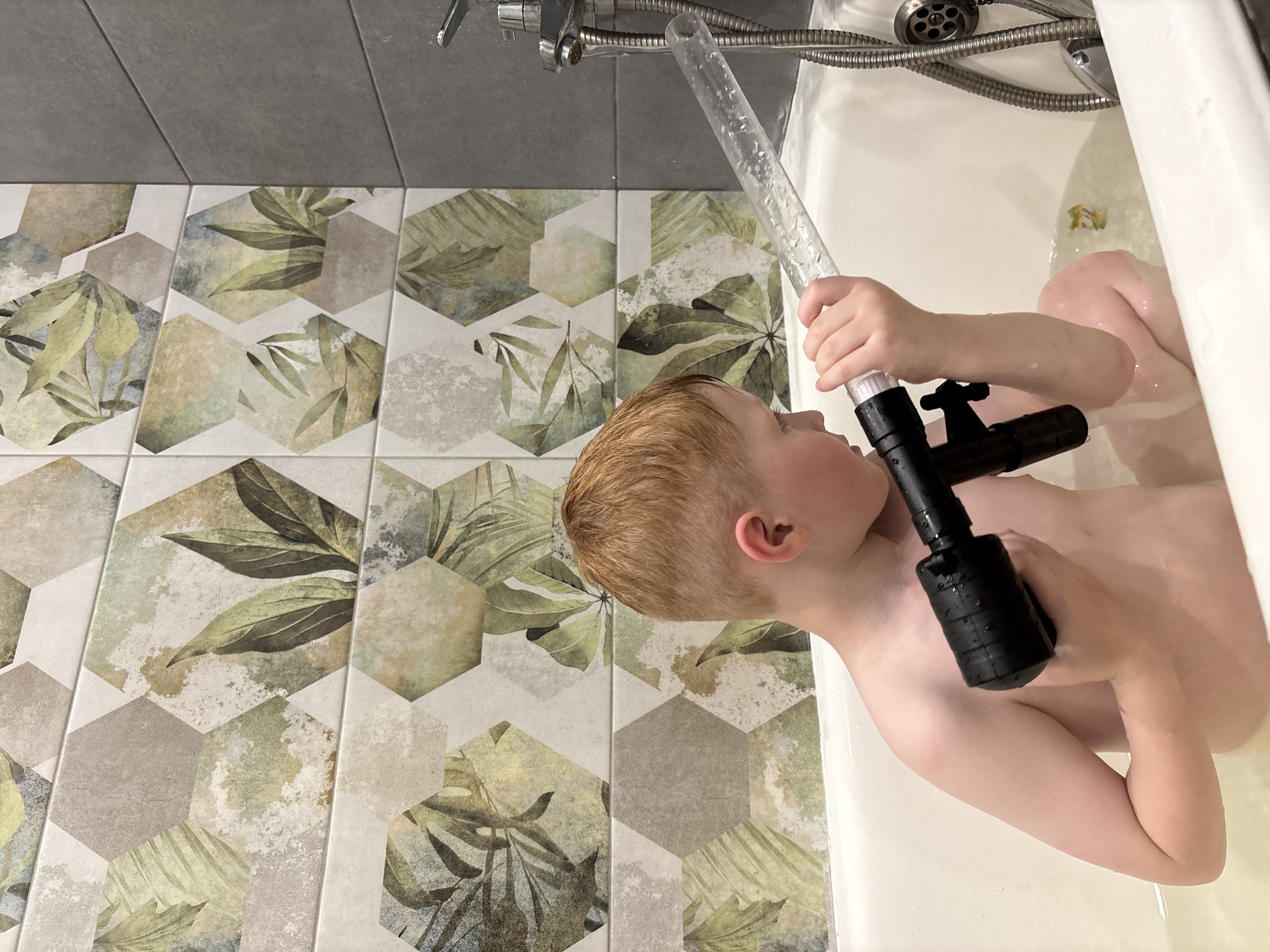 The image shows a young boy, likely between the ages of 4-6, in a bathtub filled with water. He has light brown hair and is looking to his right, holding a clear plastic tube with a black handle attached to one end. The tube appears to be connected to a shower head, from which water is flowing into the bathtub. The boy is partially submerged in the water, and his skin appears wet. He is holding the tube with his right hand.
The setting is a bathroom with tiled walls. The tiles have a geometric pattern featuring hexagonal shapes with botanical designs in shades of green, beige, and grey. The floor is also tiled with large grey tiles. The bathtub itself is white. A shower faucet with a silver hose is visible in the upper part of the image. The lighting suggests it is daytime, though no windows are visible to confirm the exact time. The boy's expression is difficult to discern, but he appears engaged in playing with the water. The overall mood is one of playful exploration and bath time fun. There is no visible text in the image. The location context is Izhevsk, Russia.