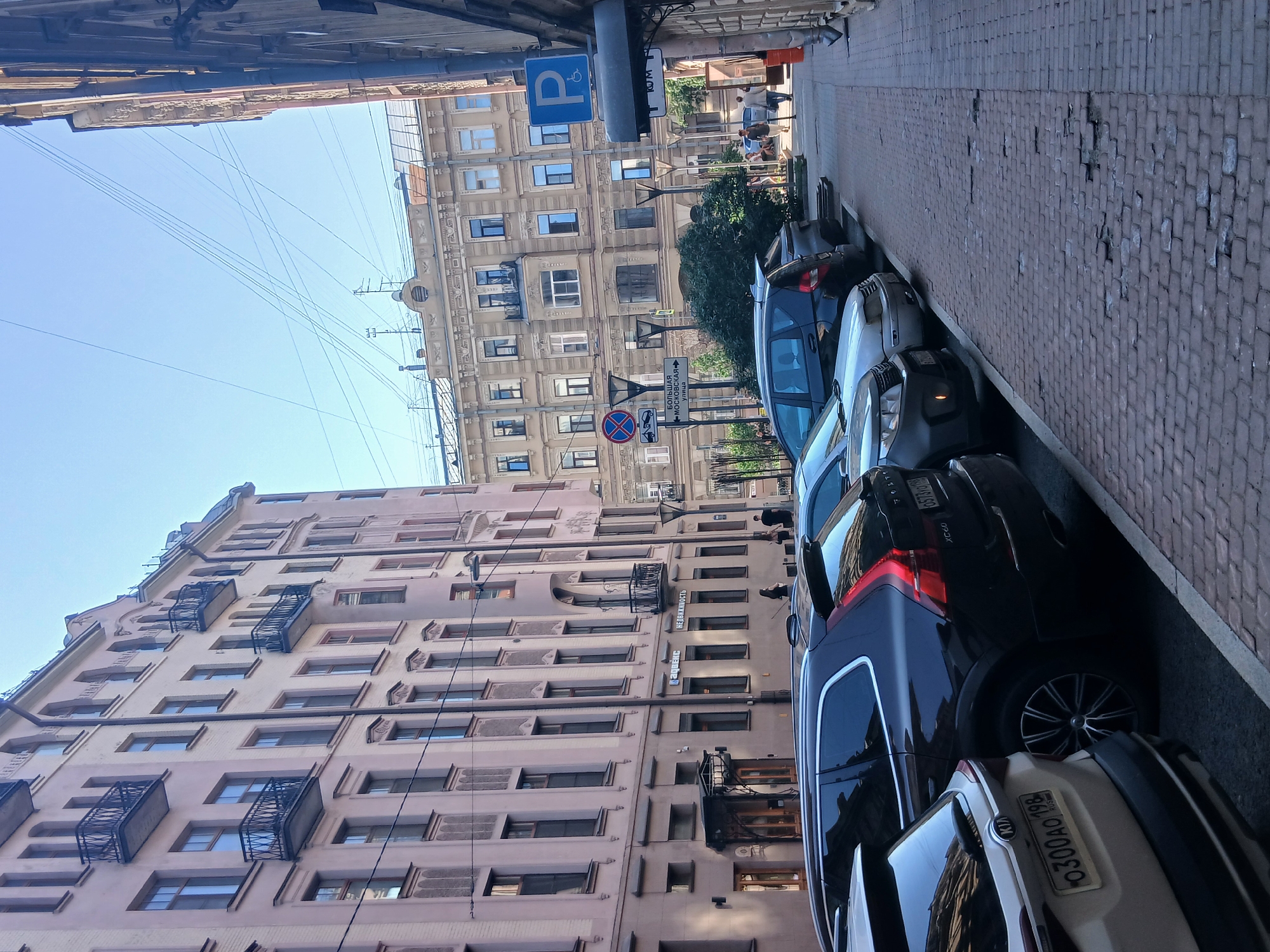 The image depicts a street scene in an urban environment, with buildings lining one side of the street and parked cars on the other. The perspective is from street level, looking down the street. The buildings are multi-story structures with a mix of architectural styles, suggesting an older city. One building on the left is a pale pink color with numerous windows and balconies. The building opposite is lighter in color and has more ornate detailing. The street itself is paved with bricks, and there is a sidewalk.
Several cars are parked along the side of the street. The cars are modern and of various colors, including black, blue, and white. Some of the cars have license plates visible, with Russian characters and numbers, such as "0300AO 198 RUS".
There are street signs visible, including a blue sign with a white wheelchair symbol, indicating a disabled parking spot. Another sign shows a red circle with a diagonal line across it, and a car symbol, likely a parking restriction sign. Further down the street, another sign has Russian text and arrows, indicating directions to "БОЛЬШАЯ МОСКОВСКАЯ".
In the background, there are a few people visible walking on the sidewalk, but they are not the focus of the image. The sky is clear and blue, suggesting a sunny day. The overall impression is a typical urban street scene on a clear day. The image is taken with a vertical orientation.