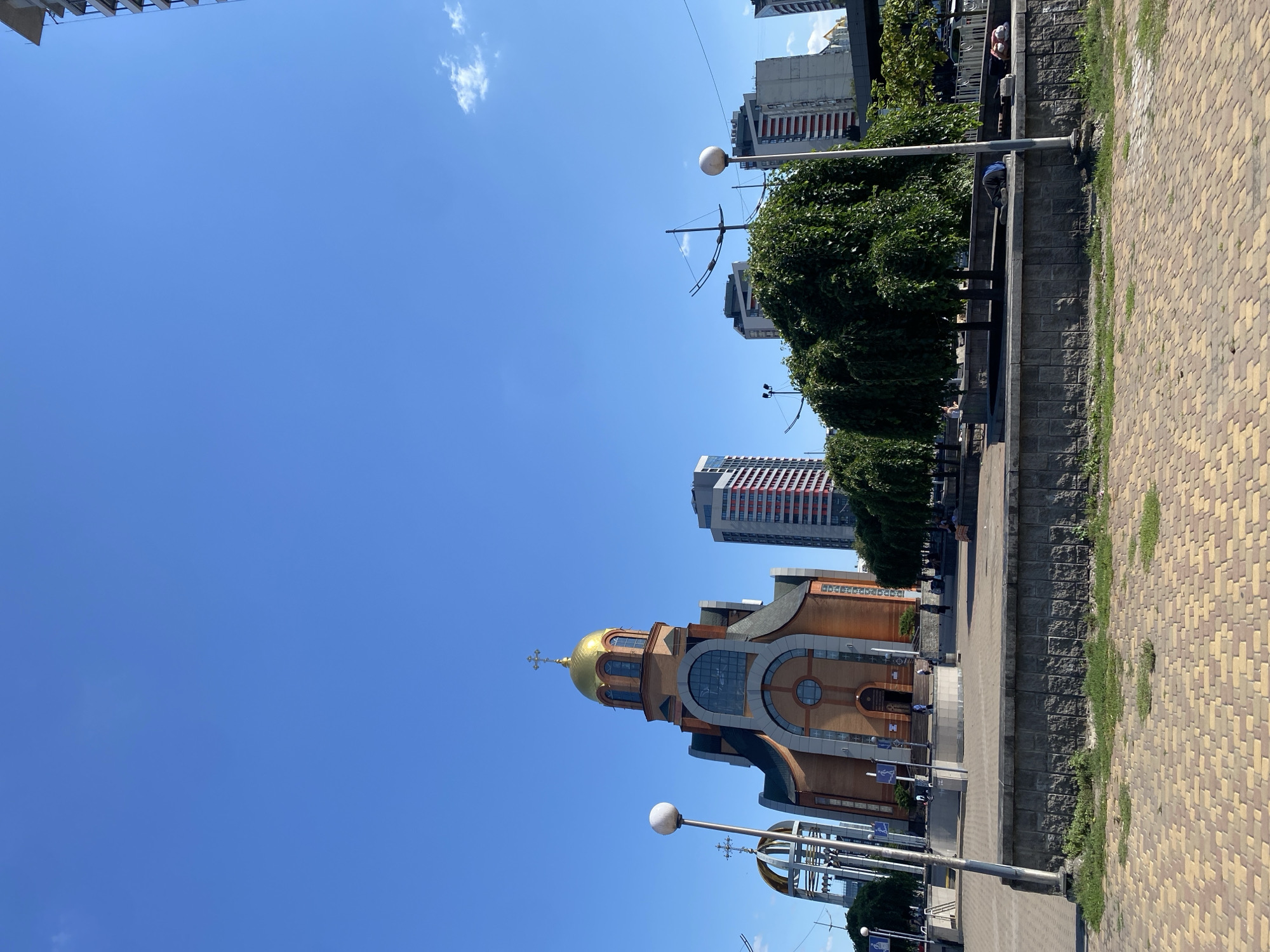 The image shows a street-level view of a plaza with a church on the left and several modern high-rise buildings in the background under a clear blue sky. The church, with a prominent golden dome and cross, is made of red brick and features arched windows. In front of the church, there is a paved walkway with a low stone wall. Several trees with dense green foliage are planted behind the wall, casting shadows. A lamppost stands near the center of the foreground. On the right side of the image, several people are seen sitting or lying down near the wall, some appearing to be sleeping. In the background, the tall buildings have a mix of grey, white, and red facades. The ground in the immediate foreground is paved with interlocking bricks in a yellow and brown pattern.