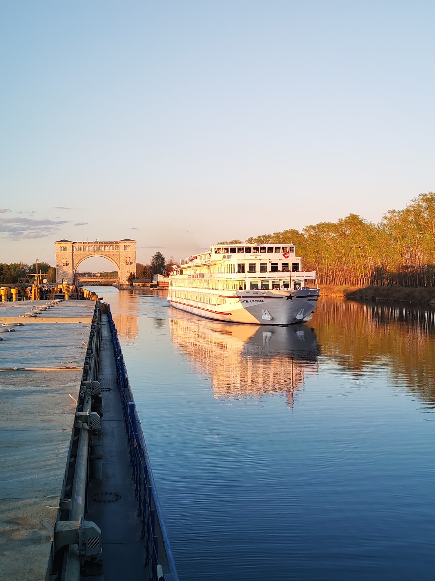A river cruise ship, the "Damian Korotkov," navigates a canal near Myshkin, Russia, as evidenced by the distinctive arched bridge structure. The ship, painted in white with multiple decks and red accents, displays passengers along its deck. Sunlight reflects off the vessel's hull and the calm water. The surrounding landscape includes a line of trees with autumn foliage along the canal bank. Along the canal bank, dock infrastructure and equipment for ships is present.
