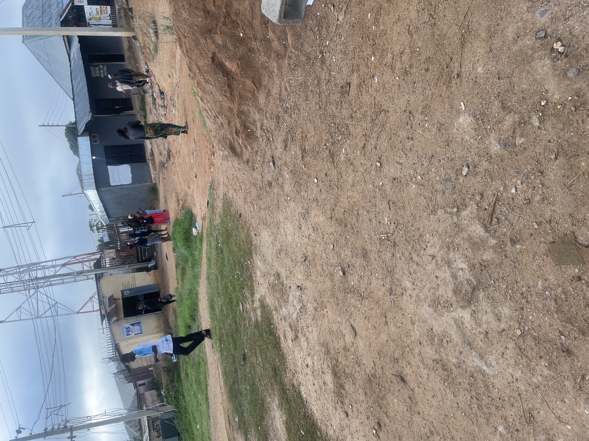 The image depicts an outdoor scene in Rafin Par, Nigeria, likely a commercial or residential area with a dirt road and scattered buildings. In the foreground, a dirt path dominates the lower portion of the frame, with patches of dry grass and scattered debris. A large pile of brown soil or sand is situated on the right side of the image.
Several individuals are present in the middle ground. On the left, a young man in a white shirt with blue accents and black pants walks away from the camera, holding a document. He appears to be moving along the edge of a grassy area. Behind him, a small building with a "POS" sign is visible.
Further into the scene, a group of people are gathered near a cluster of structures. One individual in the center wears a colorful patterned skirt and a dark top. To their right, two men are visible, one holding a shovel. Other figures are also seen milling about in front of the buildings, which appear to be shops or small businesses. One building has signage that includes "SUGAR RUSH, BAKING AND PASTRIES."
The background features a large telecommunications tower, electrical poles and wires, and more buildings with corrugated metal roofs. The sky is overcast. The overall environment suggests a developing or semi-urban area with a mix of commercial activity and infrastructure.