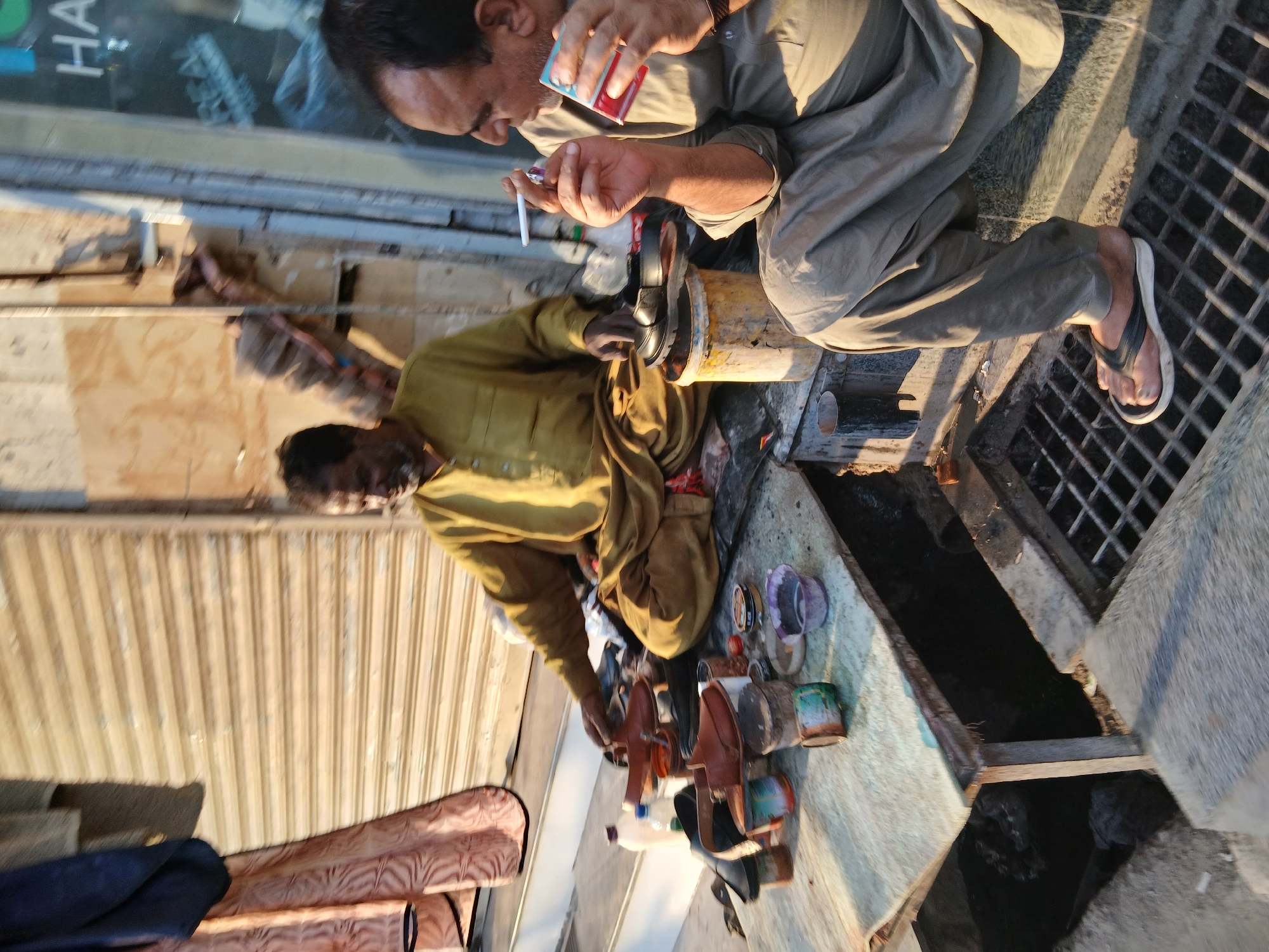 A street scene in Quetta City Tehsil, Pakistan, shows two men near a street-level drain. One man in a grey shirt is sitting, smoking, and holding a red and blue pack of cigarettes. The second man, wearing a tan, long-sleeved shirt, is working with shoes and various supplies on a surface next to the drain. There are assorted containers and bottles around him. A roll-up door and a quilted object are in the background.
