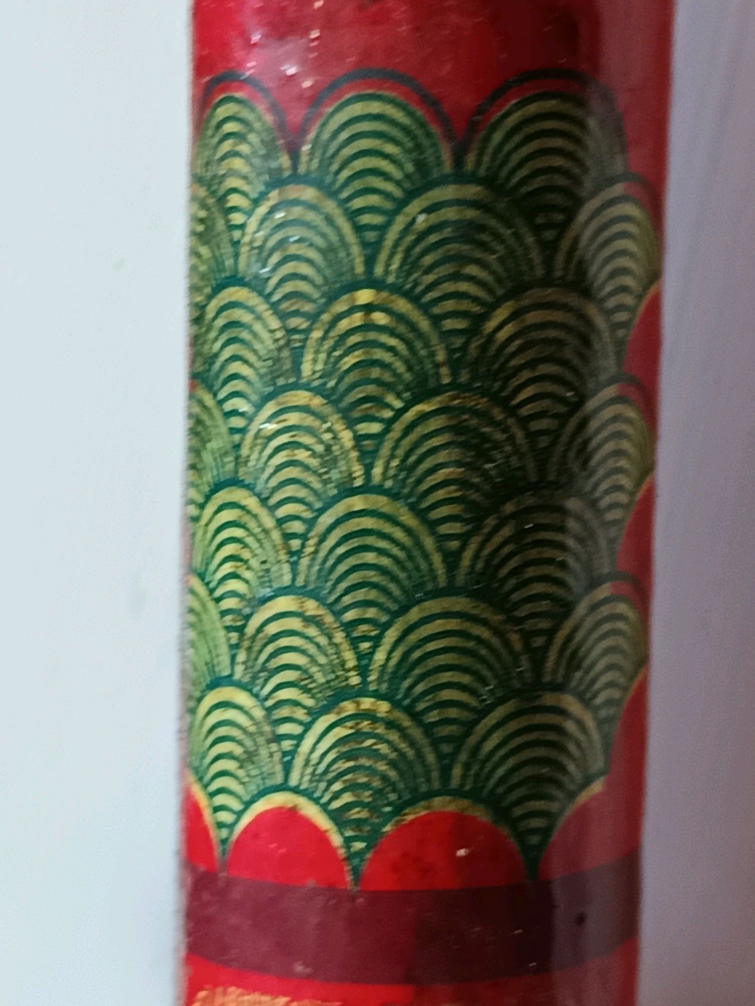 The image is a close-up of a red cylindrical object decorated with a pattern of green and yellow layered semicircles, resembling scales or waves. The pattern starts from the bottom and extends upwards. There are also red semi-circular shapes interspersed with the patterned area. The object is against a light-colored background, possibly a wall. The lighting is somewhat diffuse, with some highlights visible on the surface of the object. The scene is static, and no people or actions are depicted. The location in Gunung Sekar, Indonesia, is not visually represented in the image itself.