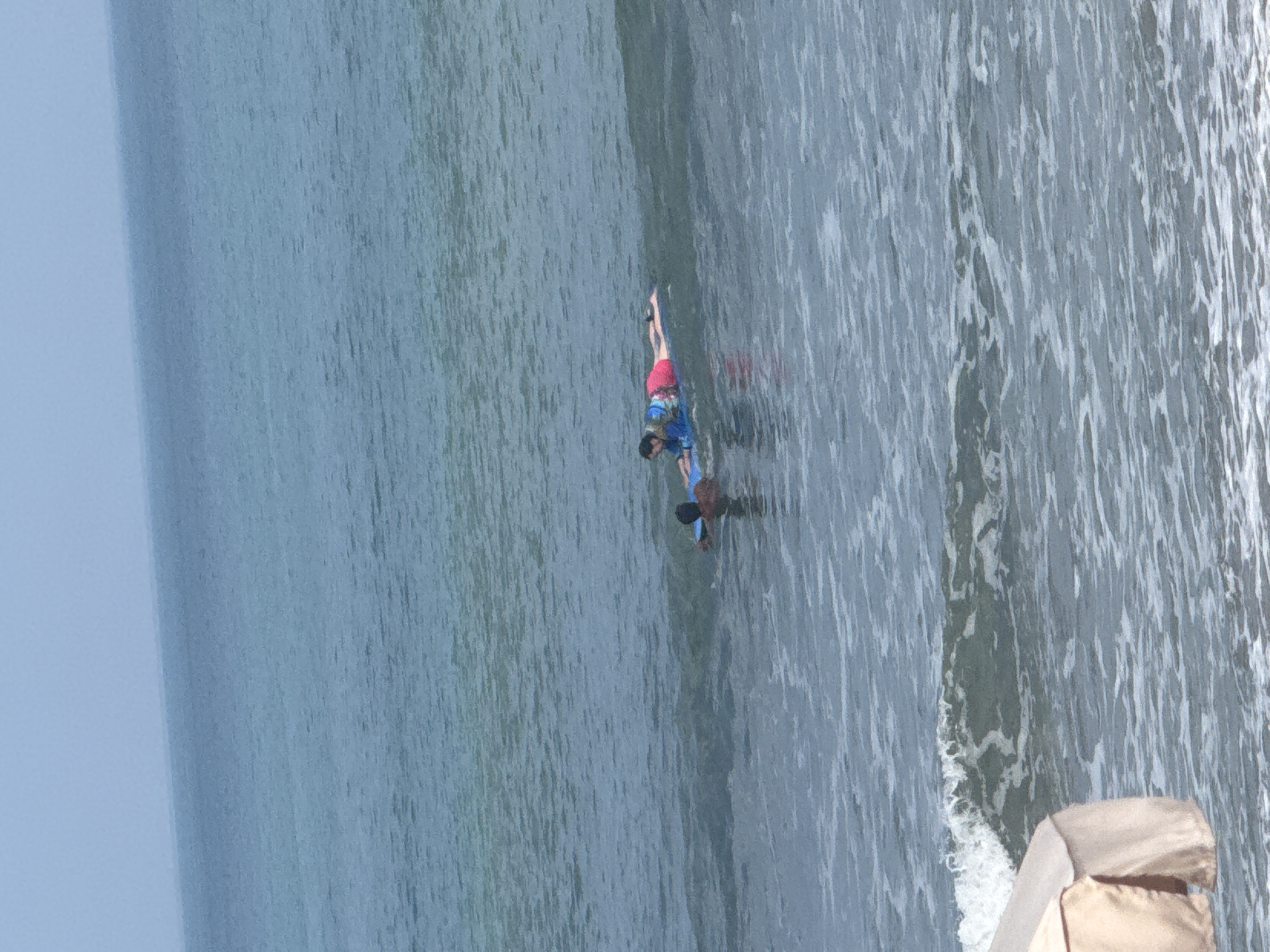 An aerial shot captures a calm ocean scene with two individuals on a surfboard near the shore of Kuta, Indonesia. The water is a tranquil blue, with gentle waves rolling towards the beach. In the foreground, the corner of a beige beach umbrella is visible, hinting at a nearby beachside relaxation area. The two people, positioned on a blue surfboard, are lying down, seemingly enjoying the water. One individual is closer to the front of the surfboard, while the other is situated further back. Their attire suggests casual beachwear suitable for a sunny day. The overall atmosphere is one of leisure and enjoyment of a serene beach environment in Kuta.