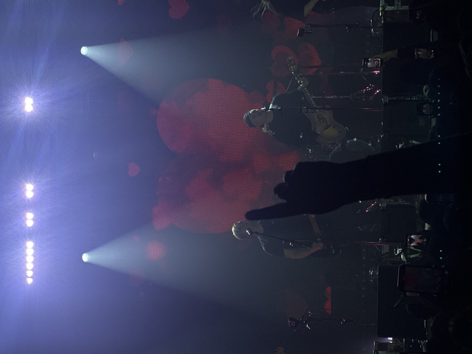 The image depicts a live music concert from the perspective of an audience member. The stage is illuminated by multiple overhead white and blue spotlights, casting bright beams through a hazy atmosphere. A large LED screen behind the performers displays a pattern of large, diffused reddish-pink heart shapes.
Two male performers are prominent on stage. On the left, a man with short, light hair, wearing a dark t-shirt, plays a dark electric guitar near a microphone stand. To his right, a second man, wearing a dark long-sleeved shirt and a dark head covering, plays a light-colored electric guitar while standing at a microphone. Additional stage equipment, including microphone stands, amplifiers, and components of a drum kit, are visible.
In the foreground, a silhouetted right arm extends upwards from the audience, forming a "rock on" hand gesture with the index and pinky fingers extended. Below this arm, multiple illuminated smartphones are visible, held aloft by audience members, presumably recording or photographing the event.
The image provides no discernible visual information to specify its precise location within the city of Izhevsk, Russia.