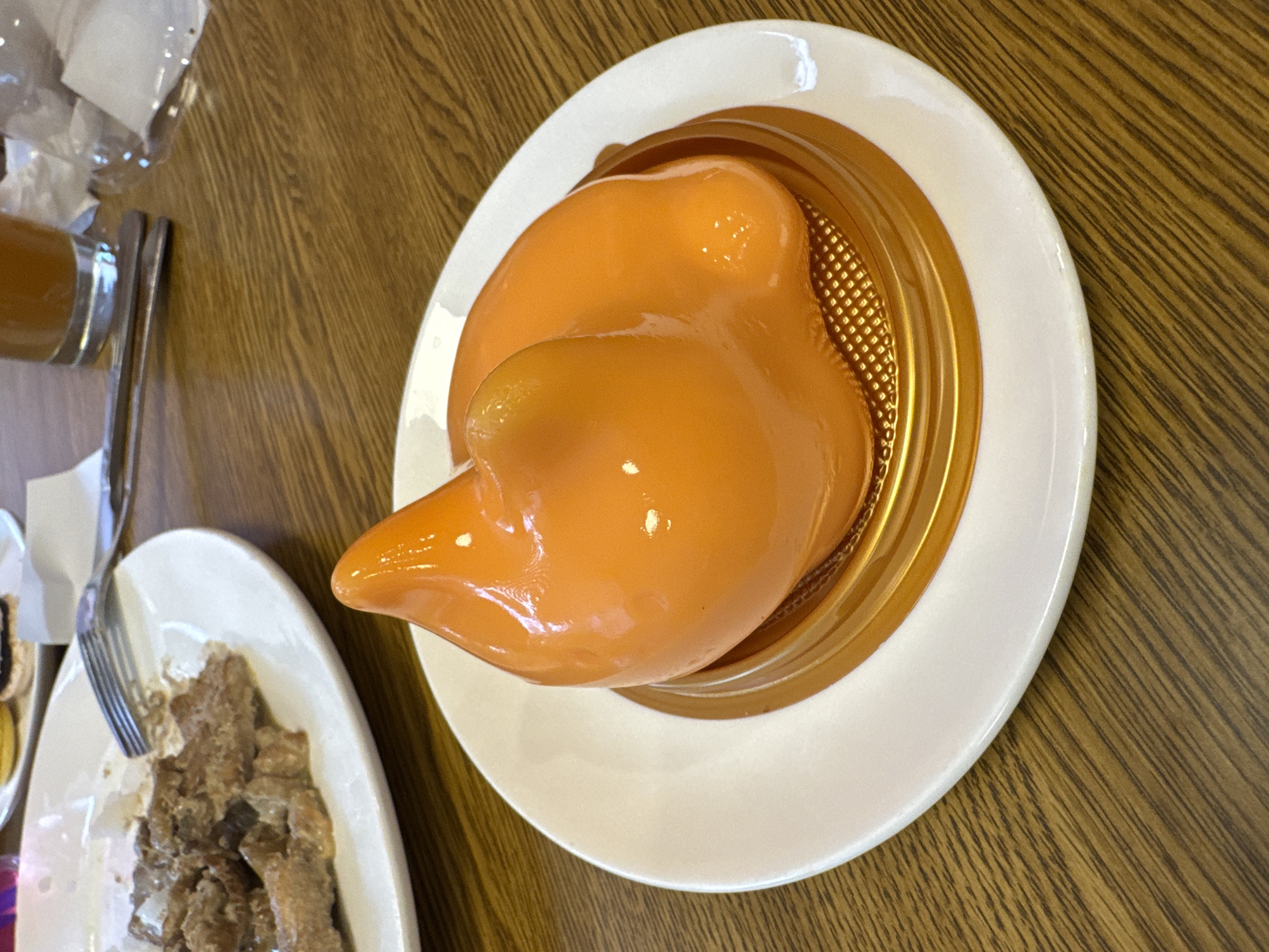 An orange, glossy, dome-shaped dessert is presented on a white plate with a gold base, resting on a wooden table. To the left, a white plate holds a portion of a savory dish, likely meat, with a fork resting on it. In the background, a glass containing a beverage is visible, along with crumpled napkins and utensils. The scene suggests a meal at a cafe or restaurant in Oskemen, Kazakhstan.