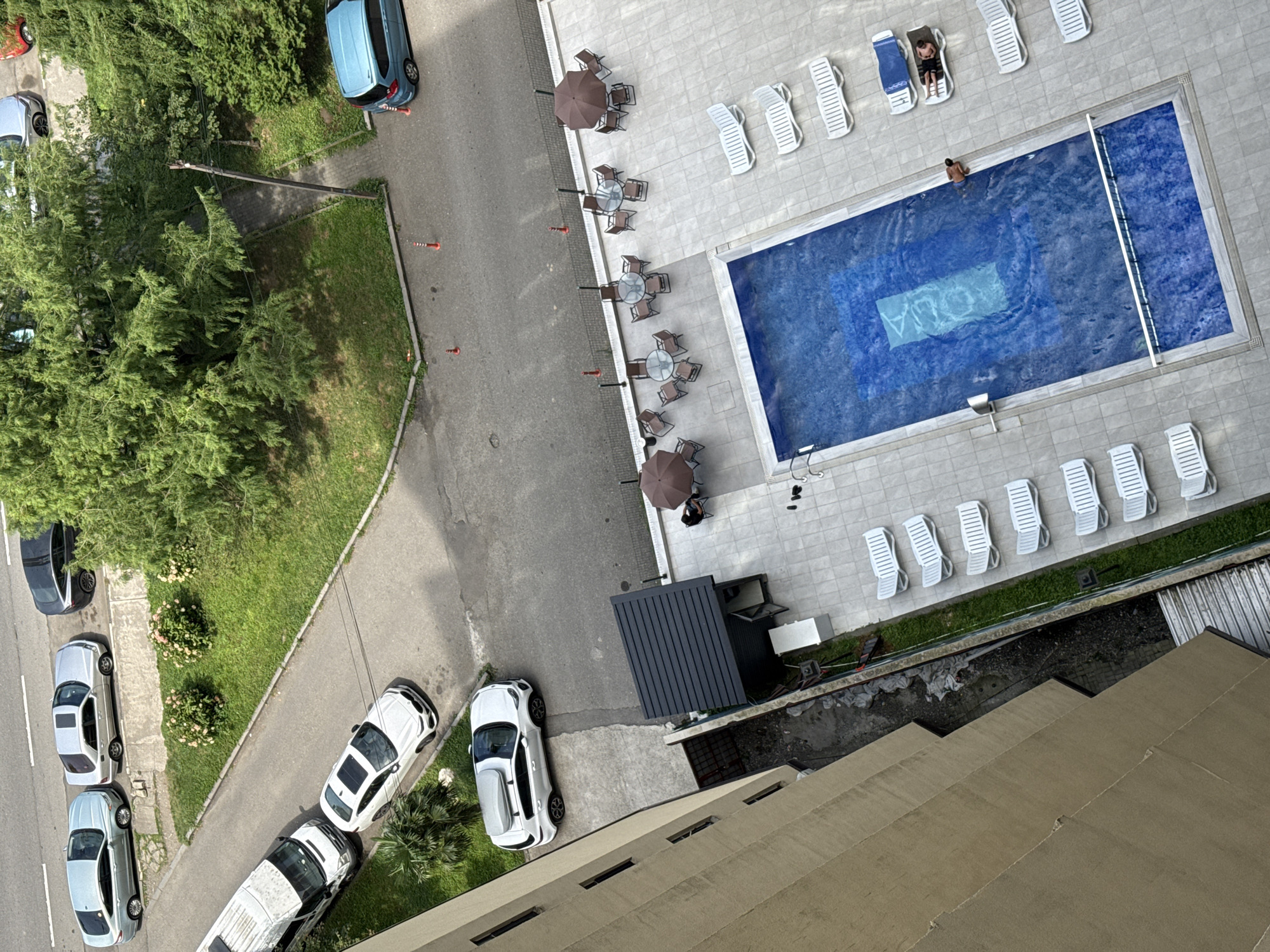 An aerial, high-angle, full shot shows an outdoor swimming pool area adjacent to a building. The pool is rectangular with blue tiles and a lighter blue rectangle in the center. A man is swimming in the pool. Around the pool, there are white lounge chairs, some occupied by people. There are also several tables with chairs and umbrellas set up on a tiled patio. To the left of the pool area is a light brown, multi-story building. To the right of the pool area is a paved road with several cars parked along the curb. Green trees and grass are visible in the background, and a sidewalk runs along the edge of the road. The scene appears to be in a residential or resort area in Batumi, Georgia.