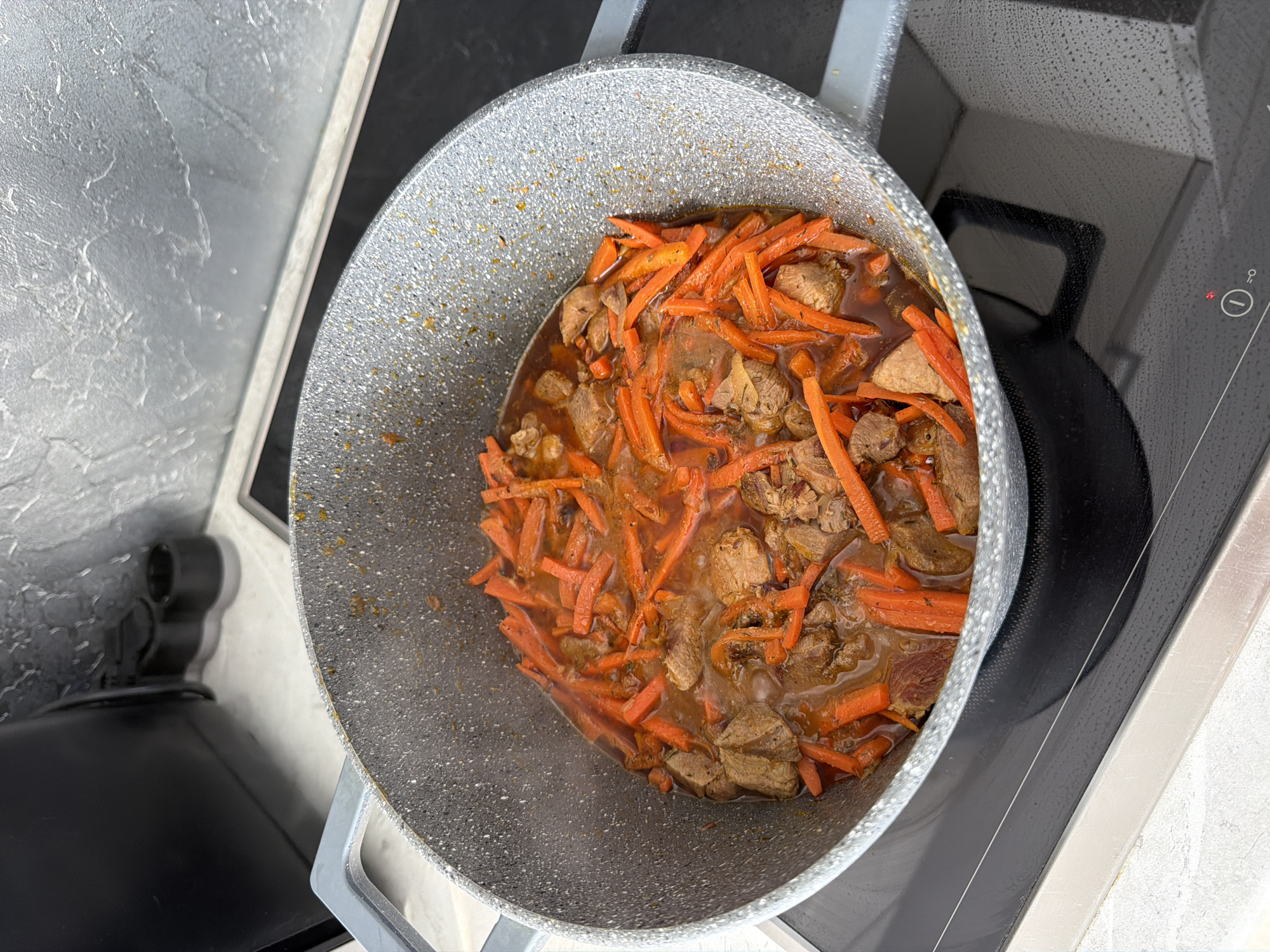 A close-up, overhead shot shows a grey speckled pot filled with a steaming stew of meat and julienned carrots. The stew is a rich brown color with visible pieces of browned meat and bright orange carrot strips. The pot is resting on a black induction stovetop in a kitchen. The background features a textured grey wall and a black appliance. There are no people or animals visible in the image. The scene is set in a kitchen, likely in a residential setting.