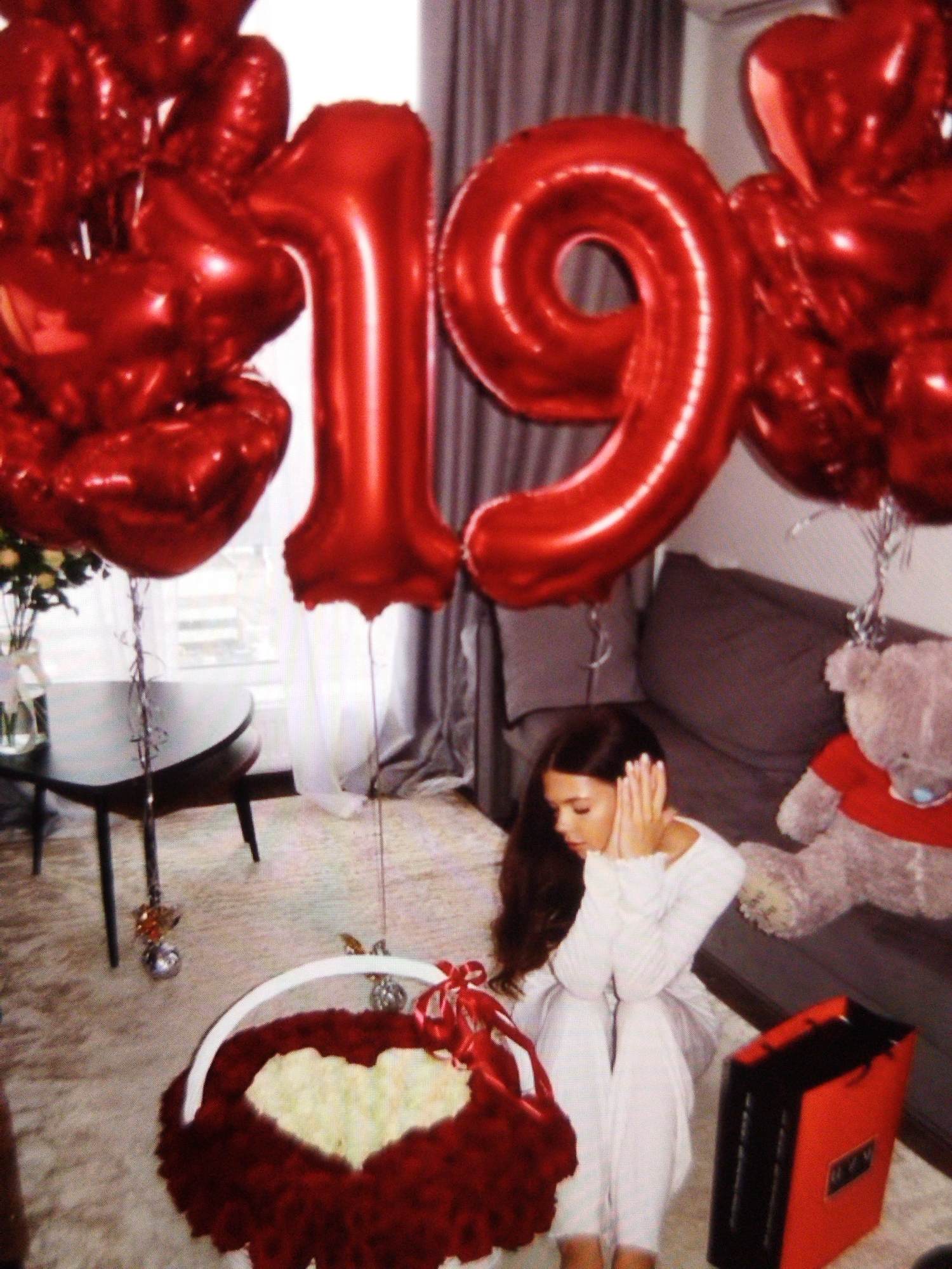 In this medium shot, a young woman with long, dark hair is sitting on the floor, surrounded by red balloons that form the number "19". She is wearing a white, long-sleeved top and white pants, and her hands are covering her ears. The woman appears distressed or overwhelmed.
In the foreground, a large basket overflowing with white roses is placed on the floor. The basket is decorated with red fabric and a red ribbon. Next to the basket, a red gift bag with black accents is visible. To the right of the woman, a large teddy bear wearing a red shirt is sitting on a couch.
The scene appears to be indoors, possibly a living room, with sheer white curtains and gray drapes in the background. A round, dark table with a vase of white flowers is situated to the left. The floor is covered with a patterned rug. The lighting suggests it might be daytime. The overall mood of the image is somber, despite the festive balloons and flowers, due to the woman's posture and expression. The location is Beșalma, Moldova.