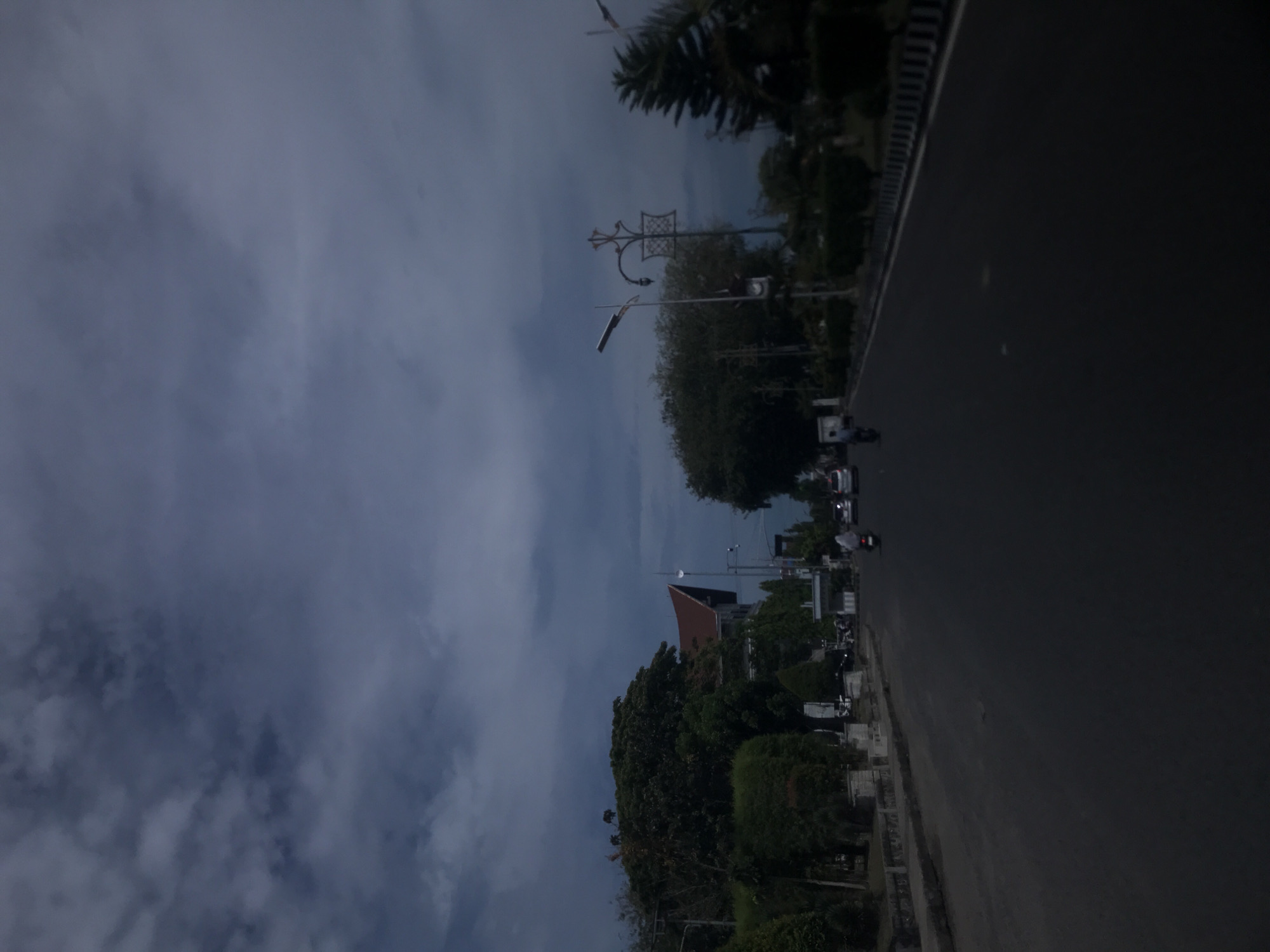 The image depicts a street scene in Lhokseumawe, Indonesia, captured on an overcast day. A road stretches into the distance, lined by sidewalks and trees on one side. Some vehicles, including cars and motorcycles, are visible on the road. In the background, a building with a distinctive roof can be seen amidst the foliage. Streetlights are placed along the road. The sky is filled with gray clouds.