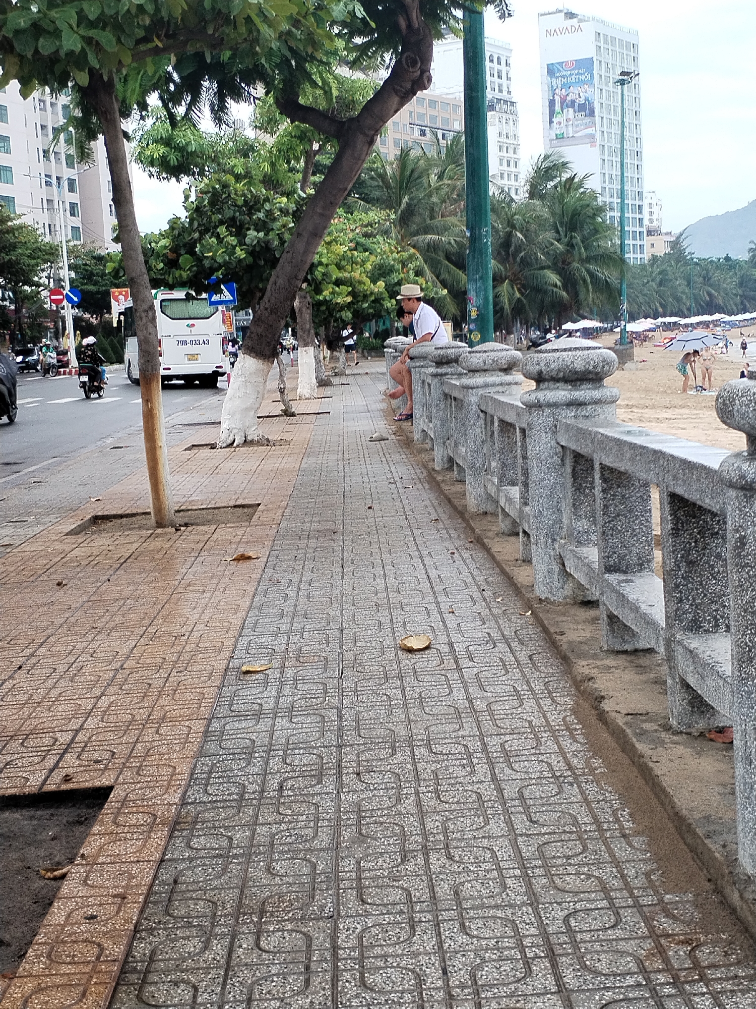 The image captures a bustling coastal scene in Bac Nha Trang Ward, Vietnam, featuring a wide patterned sidewalk, a road, and a sandy beach. In the foreground, the sidewalk is paved with two distinct patterns of grey and brown interlocking concrete tiles. A robust grey concrete railing separates the sidewalk from the beach.
On the beach, multiple white umbrellas and lounge chairs are set up, and several people are visible, including two children playing near the water's edge, suggesting leisure activities. A man in a straw hat and white shirt is seated on the railing, looking towards the beach.
Adjacent to the sidewalk is a road with a white bus bearing the license plate "79B-033.43" and several motorcycles. A row of trees with lush green foliage lines the sidewalk, some exhibiting white-painted trunks. In the background, tall, modern buildings rise above palm trees. The most prominent building displays "NAVADA" at its peak and features a large advertising billboard with Vietnamese text: "HƯƠNG VỊ HỘI TỤ LIÊN KẾT NỐI" (Flavor converges to connect), advertising a beer brand. Distant hills or mountains are visible on the horizon.
The scene takes place during the daytime under an overcast sky, indicated by the soft, diffused lighting. The atmosphere appears relaxed and lively, characteristic of a popular beach resort. Visible street signs include a blue circular "do not enter" sign and a blue square "pedestrian way" sign.