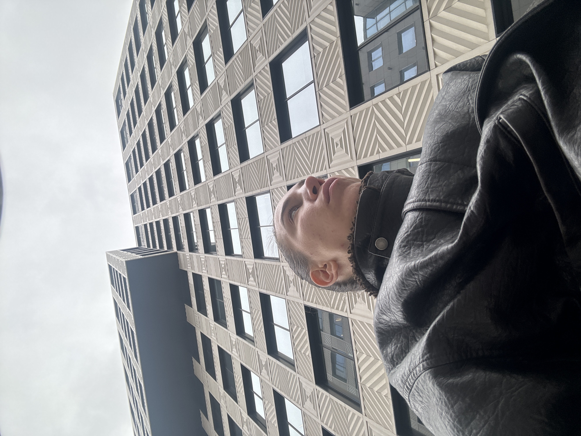 A low-angle, upward-looking shot captures a person gazing up at a modern building under an overcast sky in Moscow, Russia. The dominant subject is a tall, contemporary building characterized by a light-colored facade featuring intricate geometric patterns of diagonal lines, forming squares and diamond shapes. Dark, rectangular windows punctuate the facade, reflecting the gray sky and fragments of another distant structure. A section of the building on the left side is a solid, dark gray or blue, contrasting with the patterned main structure.
In the lower right and center of the frame, a person is depicted from the upper chest/shoulders upwards, their head tilted back as they look towards the building's height. Their profile reveals a portion of their face, including the nose, mouth, chin, ear, and one eye, with short hair visible. They are wearing a dark, textured jacket, possibly leather or a similar material, with a prominent collar that appears to have a fleece or shearling lining.
The photo is taken on a cloudy day, indicated by the uniformly light gray sky that occupies a significant portion of the left side of the image. The perspective emphasizes the towering presence of the architecture, with the building's lines converging upwards. There is no visible text in the image.