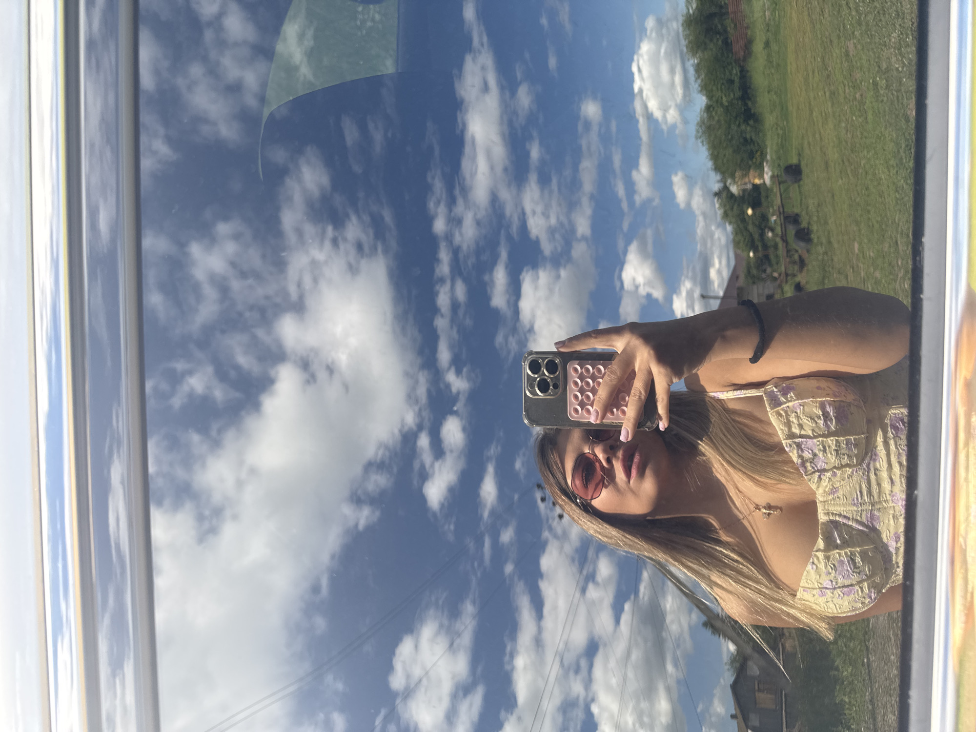 A young woman with blonde hair and pink sunglasses takes a selfie in a car mirror, reflecting a bright blue sky with fluffy white clouds. She is wearing a yellow top with a floral pattern and a gold cross necklace. Her right hand holds a smartphone with a pink pop socket attached to the back. In the background, reflected in the mirror, is a rural landscape with houses, trees, and a field. The image appears to have been taken outdoors on a sunny day.