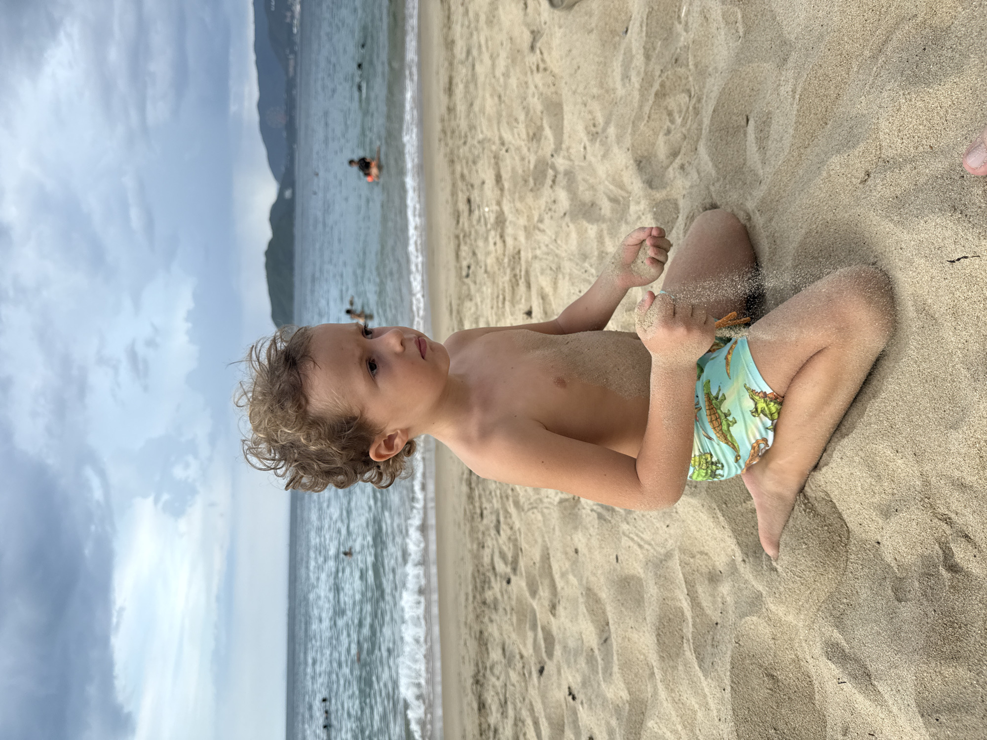 A young boy with curly brown hair sits on a sandy beach, playing with sand. He is wearing light blue swim trunks with a pattern of green and orange alligators on them. His body is partially covered in sand, suggesting he has been playing in it. He is looking off to his right with a thoughtful expression.
In the background, the ocean stretches out, with small waves breaking near the shore. There are several people in the water, some swimming and others wading. In the distance, a mountainous coastline is visible under a cloudy sky. The lighting suggests it is daytime, possibly late afternoon given the soft light and overcast conditions. The scene is set at a beach in Nha Trang, Vietnam. No text is visible in the image.