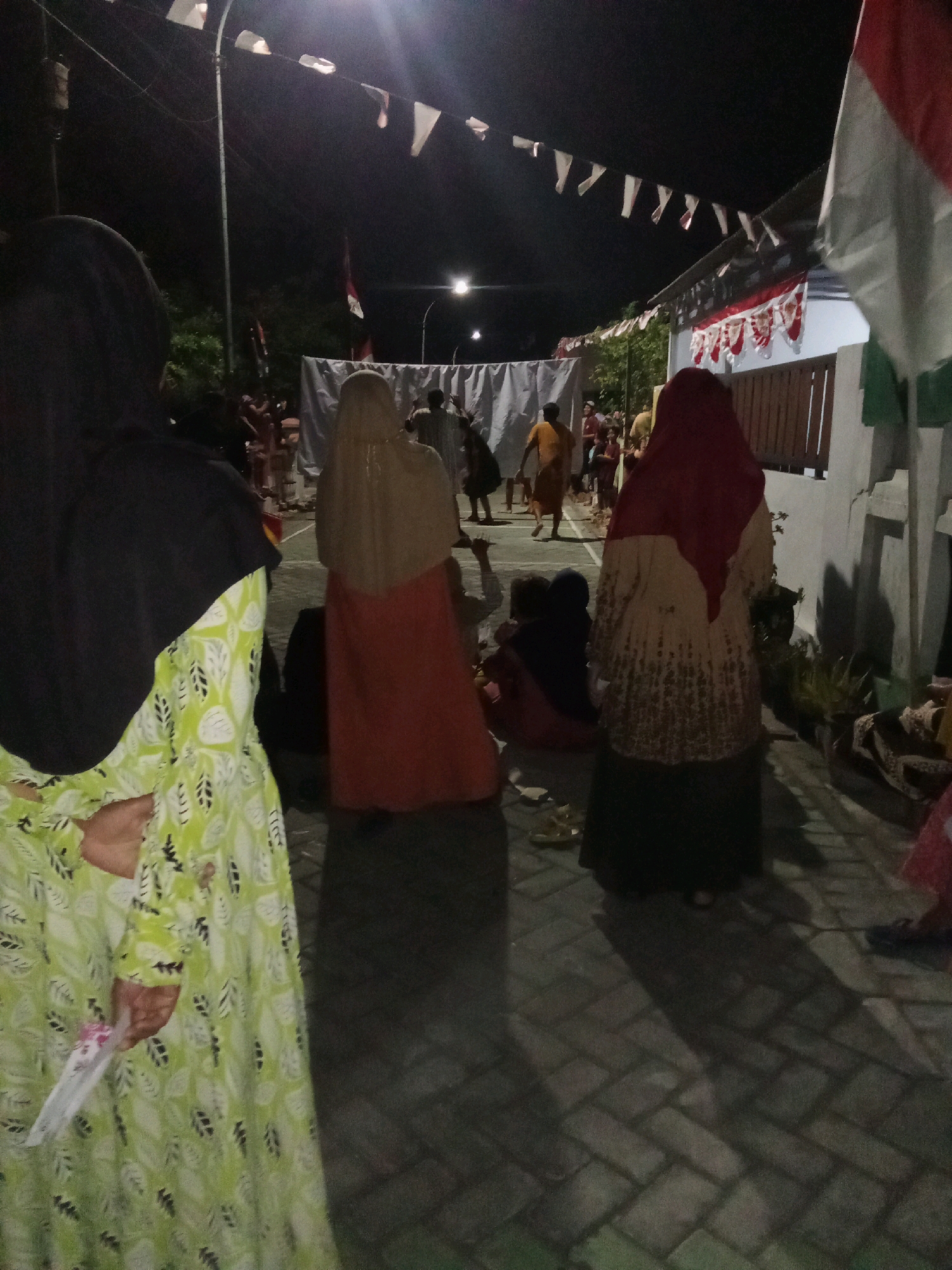 This image depicts a nighttime outdoor gathering in the city of Sidorejo, Indonesia. A group of people are seen from behind, with some individuals wearing hijabs. One person in the foreground is wearing a light green dress with a leaf pattern and a black hijab, holding a small white object. In the middle ground, several people are gathered, some seated on the ground. They appear to be watching an event or performance taking place in front of a white sheet or screen. In the background, there are strings of triangular flags, a street lamp, and a building decorated with more flags and banners. The ground is paved with interlocking bricks. The overall scene suggests a community event or celebration taking place in a residential area at night.