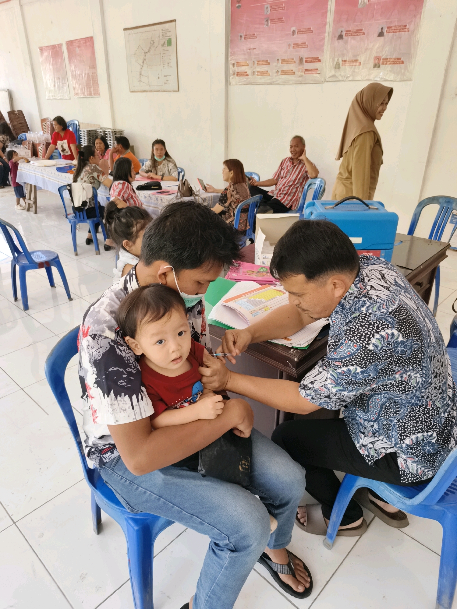 The image depicts a vaccination event at a community hall in Tombatu Dua, Indonesia. In the foreground, a man in a patterned shirt and blue jeans is holding a young child, who is wearing a red shirt. The man is wearing a light blue face mask and appears to be administering a vaccine to the child. To the right, another man in a patterned shirt is seated, looking down at a book. In the background, several people are seated at tables, some wearing masks, suggesting a crowded public gathering. The room is sparsely furnished with rows of blue plastic chairs. Posters are visible on the wall. The overall atmosphere appears to be one of organized public health activity.