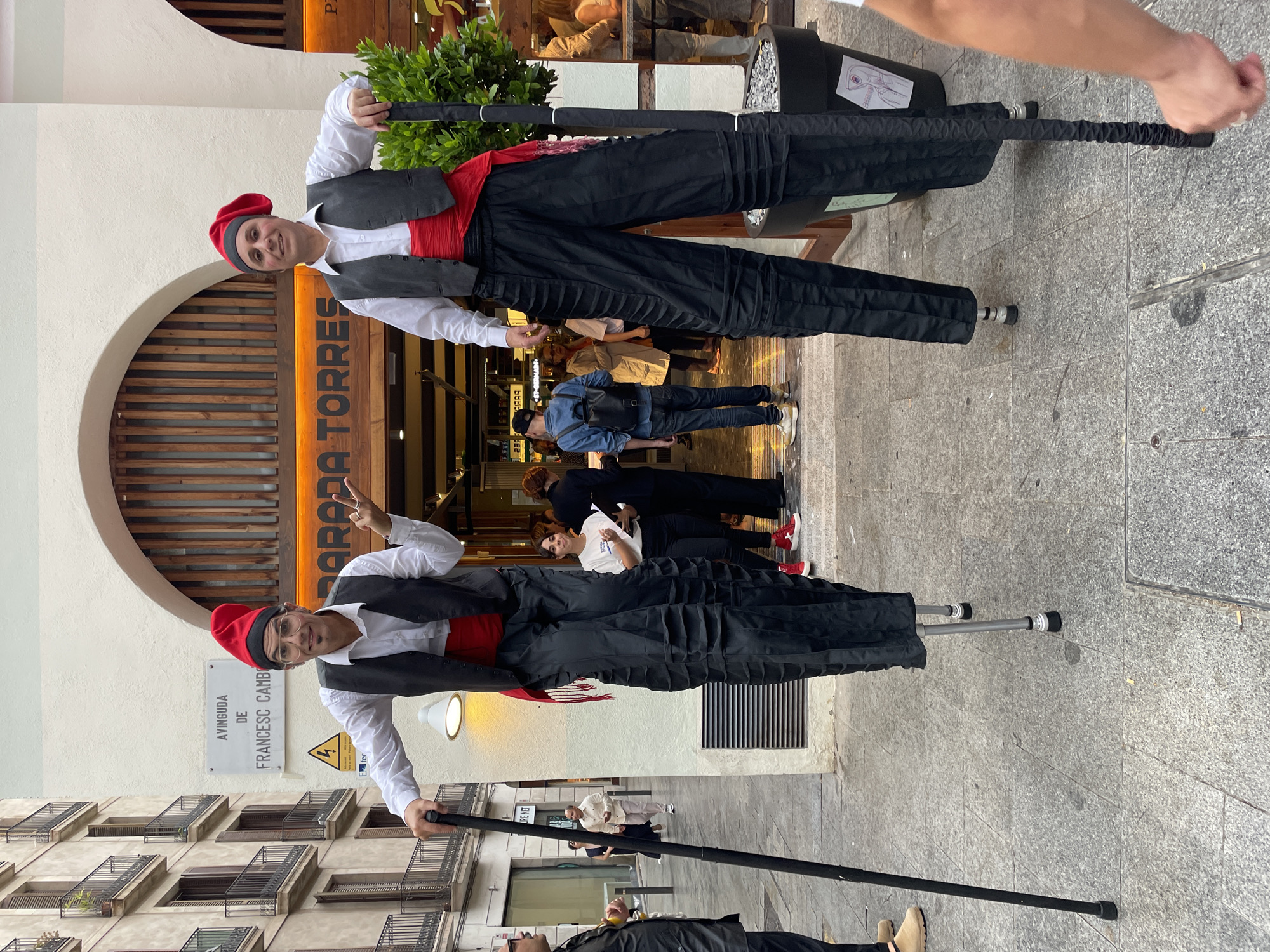 The image captures two male street performers on stilts, dressed in traditional Catalan attire, on a paved street in Barcelona. Both men wear white long-sleeved shirts, dark waistcoats, red sashes (faixes), black trousers, and red barretinas (traditional caps). The performer on the left smiles directly at the camera, while the one on the right smiles and forms a peace sign with his hand. Their black stilts elevate them significantly above the ground.
The scene unfolds during the daytime under clear weather, with natural light illuminating the street. In the background, multi-story buildings with balconies line the avenue. A shop with a distinctive wooden facade and an arched entrance is visible, displaying a partially legible sign that appears to read "AROPA TORRES" or similar. Several pedestrians can be seen in the background, including a person with a backpack.
A white street sign affixed to the building clearly indicates the location as "AVINGUDA DE FRANCESC CAMBO" and includes the word "AYUNTAMIENTO" (City Hall), pinpointing the specific area in Barcelona. Below this, a yellow triangular warning sign with a lightning bolt symbol is also visible. The ground is covered with grey stone pavement. The overall impression is one of an engaging street performance in a vibrant urban environment.