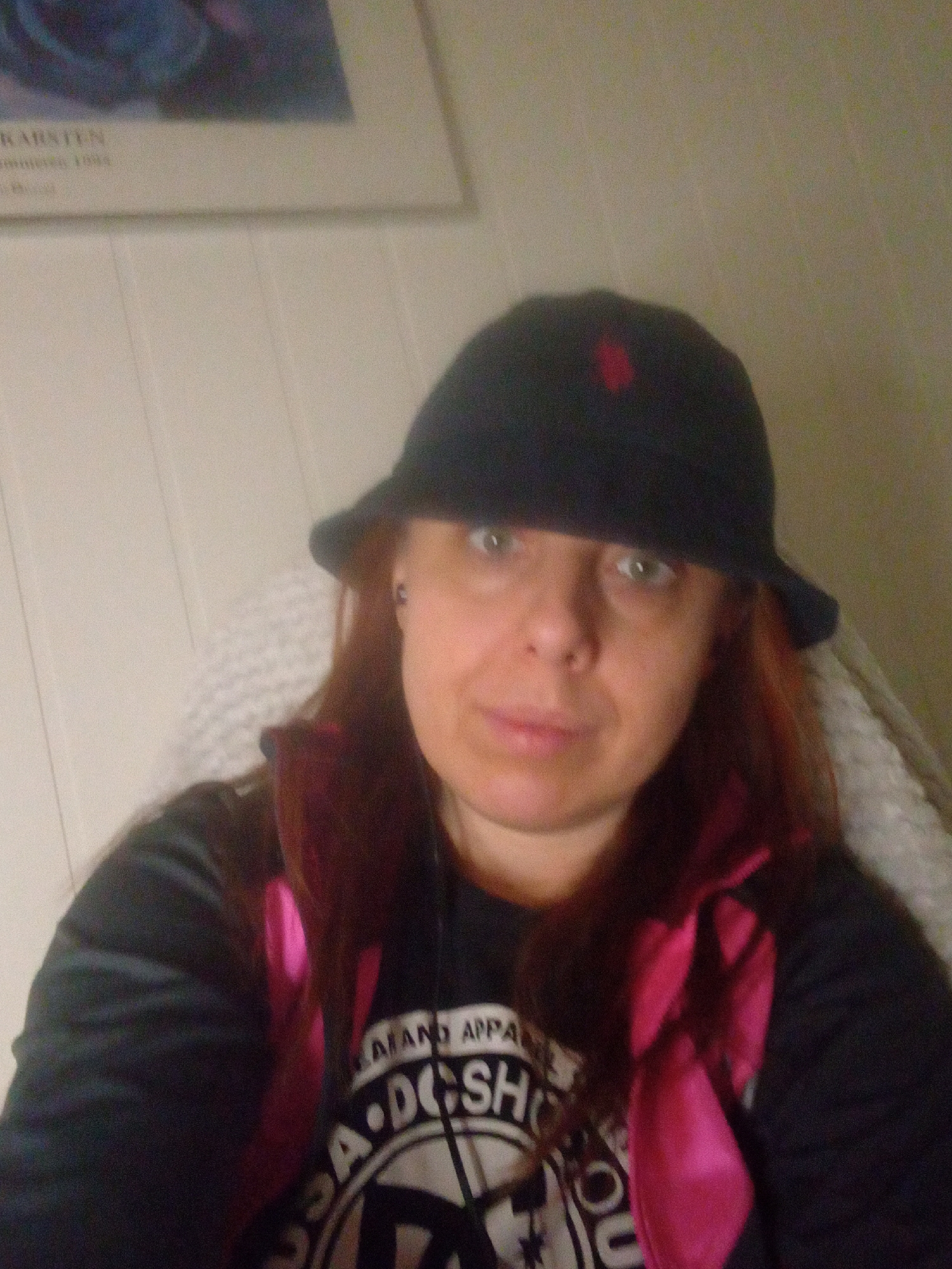 An adult female is depicted in a medium-close-up, frontal shot. She has fair skin, reddish-brown hair, and light-colored eyes. She wears a dark bucket hat with a small red emblem on the left front, covering her forehead and partially her ears. A dark earring is visible in her left ear. She has a neutral facial expression. Her attire includes a dark long-sleeved garment layered over a light-colored T-shirt featuring a white graphic design with partial text "A AND APPA" and "A.DCSHO". A prominent bright pink ribbon or strap is visible around her neck and chest. She is seated, with a light-colored, textured object (possibly a blanket or cushion) behind her right shoulder. The background features a light-colored, vertically paneled wall. In the upper left background, a framed poster is visible with faintly legible text "KARSTEN" and "maritime 1984" beneath a blurred image of blue and purple tones. The lighting appears to be frontal. The image exhibits noticeable blur and graininess. The location is an indoor setting in an unknown city.