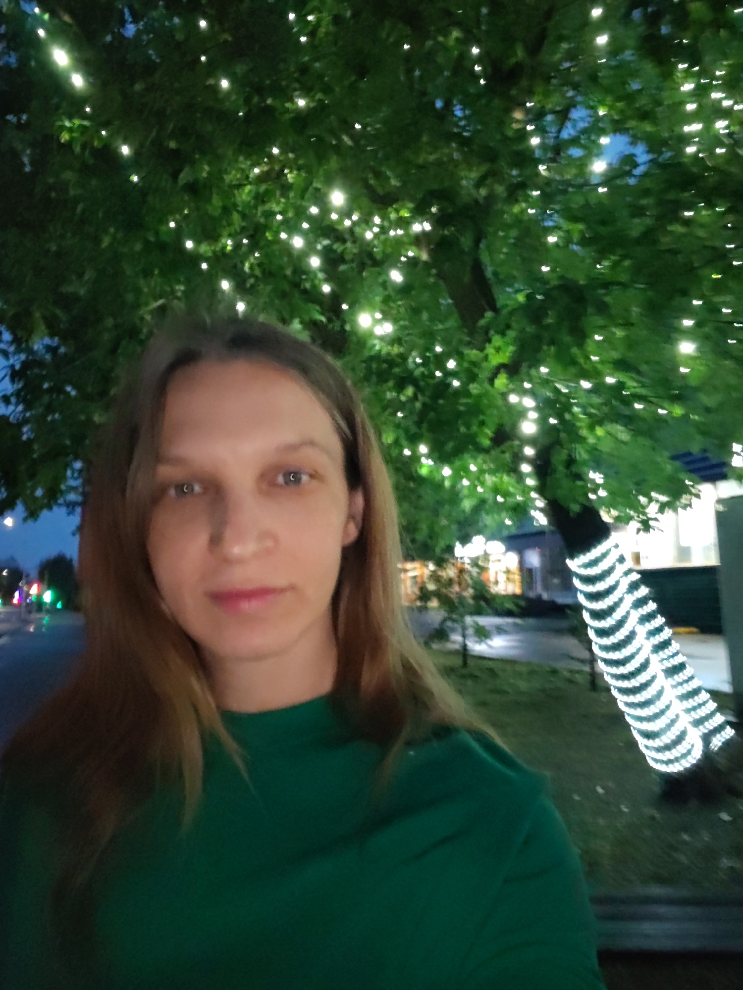 A woman is taking a selfie in front of a tree decorated with white lights. The woman has light brown hair and is wearing a green shirt. The tree is in the background, and it is nighttime. The location is identified as Unknown, Unknown.