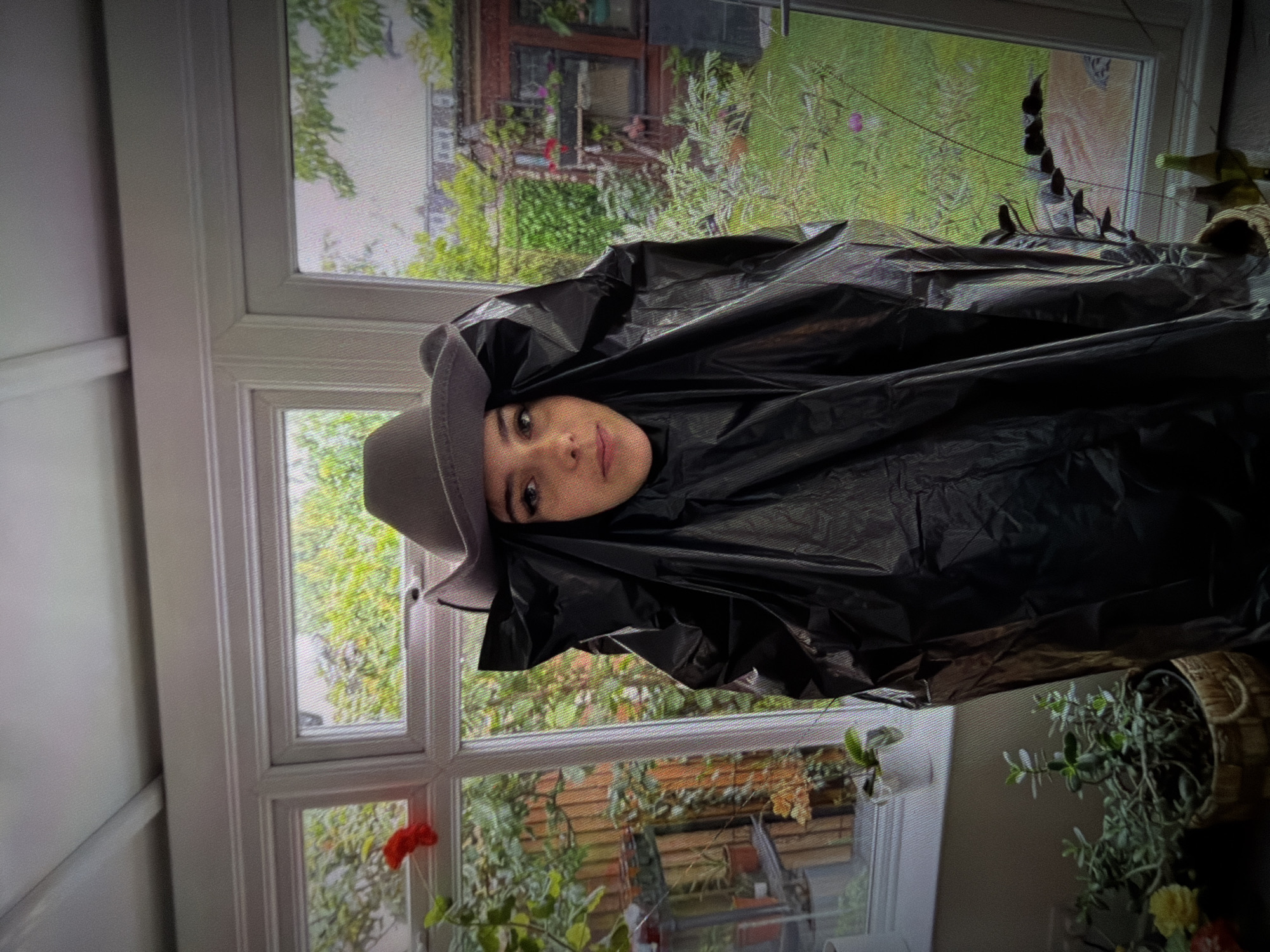 A person wearing a gray cowboy hat and a black tarp stands in front of a window. The person's face is visible, and they are looking at the camera. The tarp covers most of their body, draped over their shoulders and extending downwards. Behind them, through the window, a backyard scene is visible with green grass, trees, and a red brick structure. There are several potted plants in the foreground, some on a windowsill and others on the floor. The overall lighting suggests it is daytime.