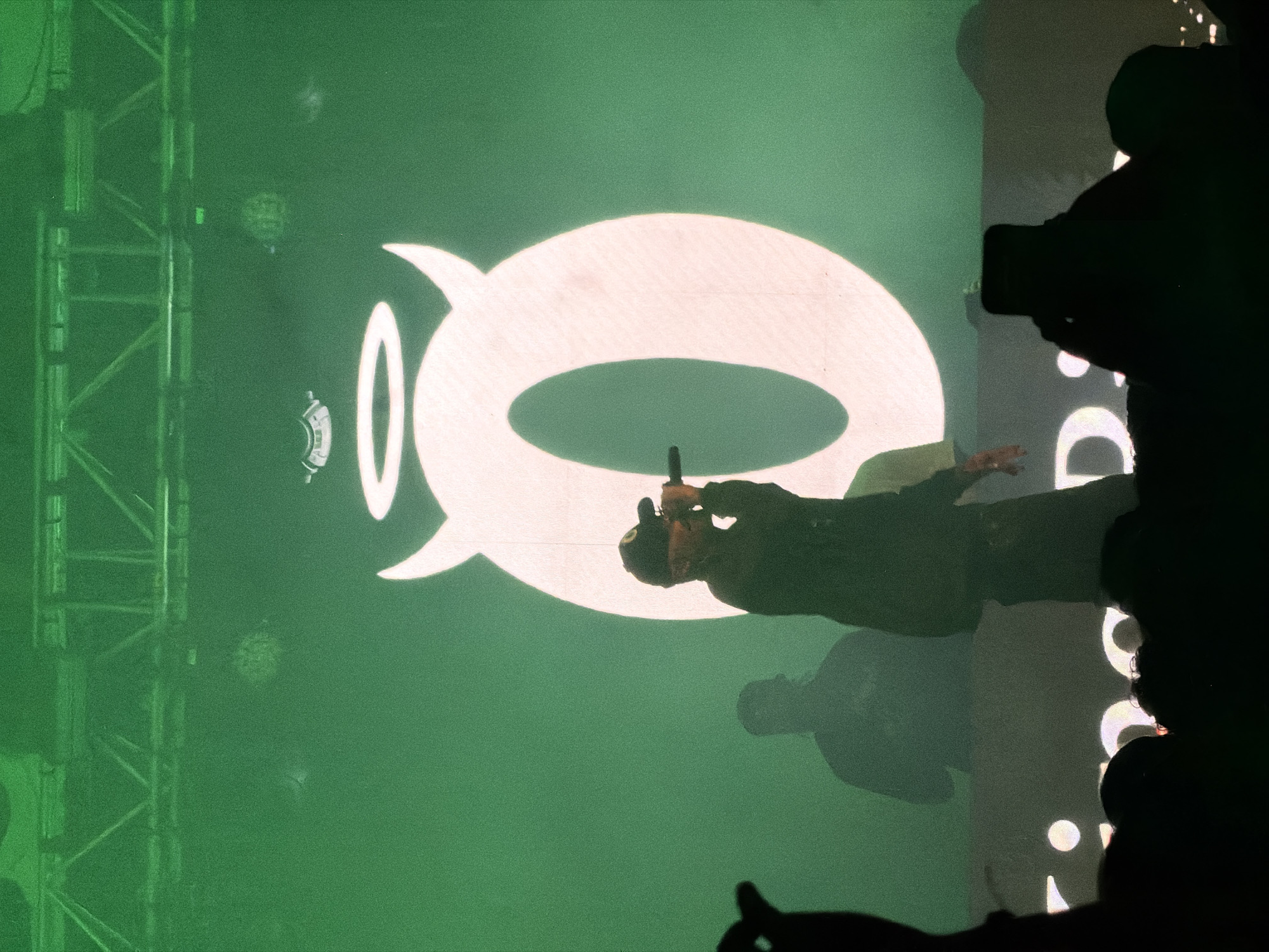 This is a vertically oriented image captured at a live performance event. The central figure is a performer, visible as a dark silhouette holding a microphone, standing on what appears to be a stage. They are illuminated from the front, casting a distinct shadow on the large green screen behind them. This screen displays a prominent, bright white, highly stylized symbol resembling the letter 'Q', with a distinctive top-left spike and a bold, hollow oval shape.
The background is dominated by this vibrant green hue from the projection. To the left of the performer, a metal truss structure is visible, suggesting stage scaffolding or lighting equipment. On the right side and extending towards the bottom of the frame, dark silhouettes of an audience can be seen, some appearing to hold up phones to capture the event. These silhouettes create a strong contrast against the brighter projected light.
The scene is lit by artificial stage lighting, giving the image a high-contrast and dramatic feel. The strong green and white colors indicate a specific lighting design for the performance. The event is taking place at night or indoors, given the reliance on artificial light. Below the performer, partial white letters on the green background are barely visible, but no clear words can be deciphered. The overall atmosphere suggests an energetic concert or show.