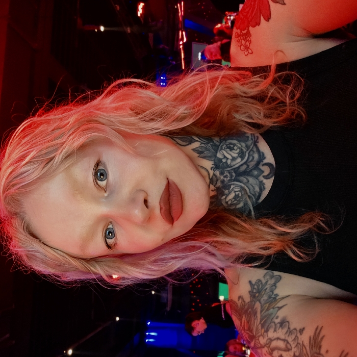 The image depicts a self-portrait of a woman in an indoor entertainment venue in Saint Petersburg, Russia. The woman has wavy, shoulder-length blonde hair, blue eyes, and a pale complexion, accentuated by dark red matte lipstick and thin or absent eyebrows. Her neck is extensively tattooed with intricate designs featuring roses, skulls, and a crescent moon, while additional floral or ornamental tattoos are visible on her shoulders and upper arms. She is wearing a plain black tank top. The background is dimly lit with artificial, colored lighting, predominantly red on the right and blue on the left, revealing blurred figures of other individuals and indistinct structural elements consistent with a nightclub or bar environment.