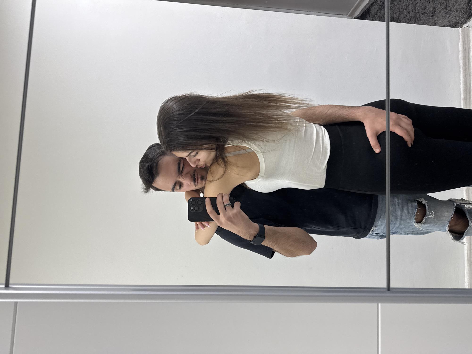 A couple is taking a selfie in a mirror. The man has dark hair and is wearing a black t-shirt and ripped blue jeans. The woman has long brown hair and is wearing a white tank top and black leggings. The man is holding a black smartphone. The location appears to be indoors, possibly a bathroom or dressing room, but the city of Sofia, Bulgaria cannot be determined from the image.