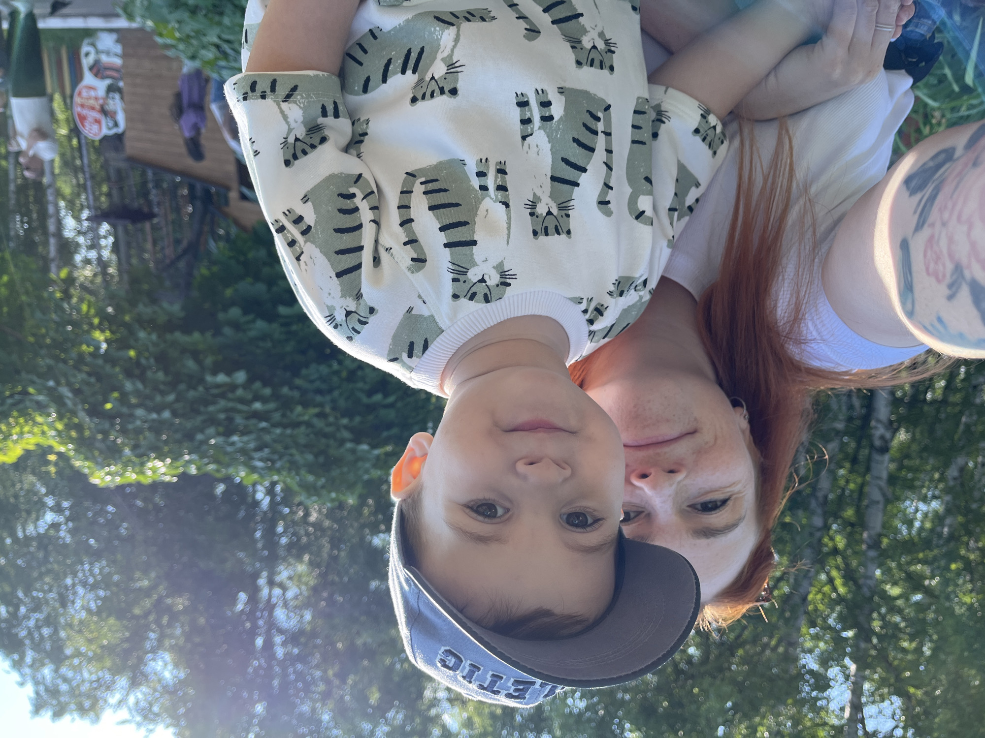 A red-haired woman with freckles and a tattoo on her left arm is taking a selfie with a young boy in a park. The boy is wearing a gray baseball cap with "ATHLETIC" written on it and a white t-shirt with a pattern of green tigers. The woman is wearing a white t-shirt. They are both smiling. The background is a lush green park with trees and sunlight filtering through the leaves. In the distance, there are some buildings and other people. The scene appears to be during the daytime, likely in the summer. This image was taken in Chaikovsky, Russia.