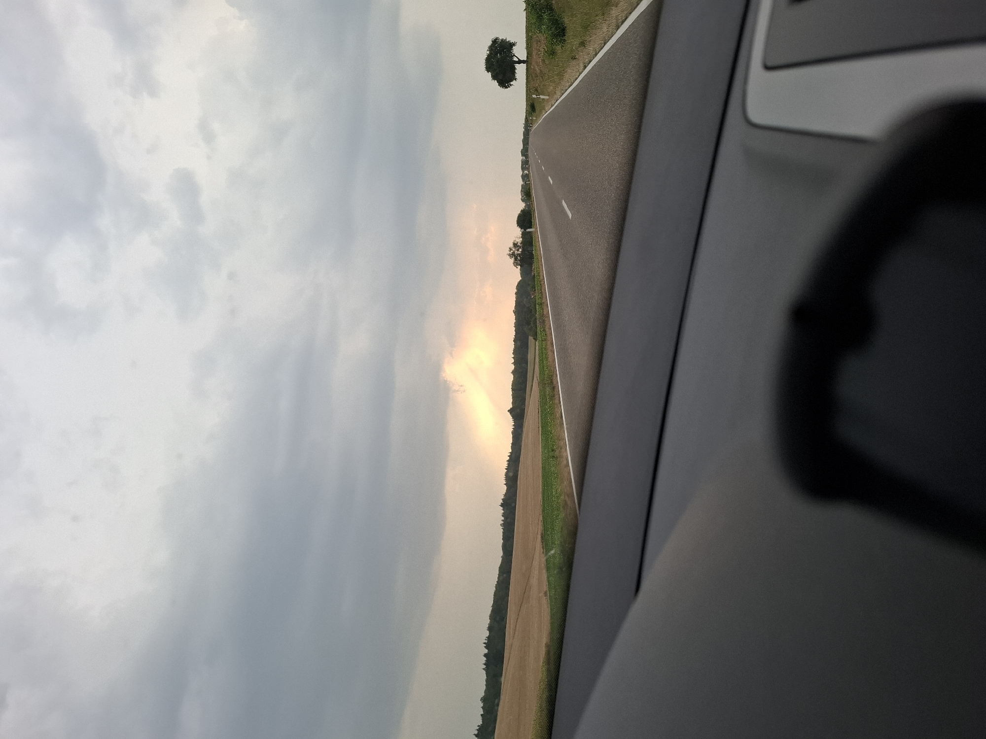 The image depicts a view from inside a vehicle looking out at a road stretching towards the horizon under a cloudy sky. The road is asphalt and has white dashed lines marking the lanes. To the left of the road are fields, one appearing to be harvested wheat and another with green crops. A line of trees borders the fields in the distance. To the right of the road, there is a grassy verge with a solitary tree. The sky is filled with heavy, gray clouds, with a patch of brighter, warmer light breaking through in the distance, possibly indicating a sunlit area or distant sunset. The foreground shows the dashboard and steering wheel of the vehicle, out of focus. The scene appears to be in a rural setting, possibly on the outskirts of Bad Rappenau, Germany, given the agricultural landscape. There are no people or other vehicles visible in the immediate vicinity.