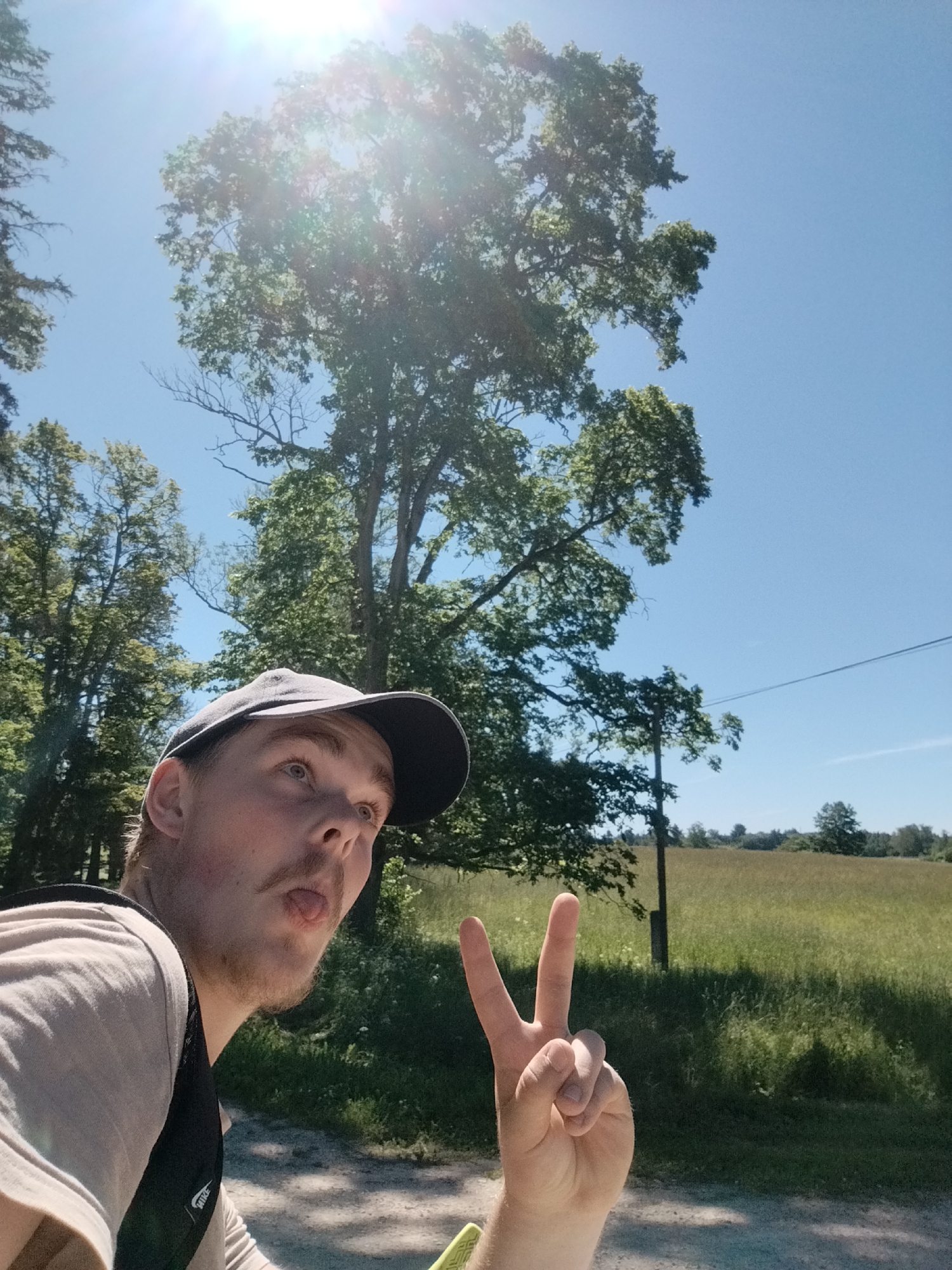 In a sun-drenched outdoor scene, a young man with a cap is pictured in a selfie-style pose. He is looking upwards and to his left, with his tongue playfully sticking out. His right hand is raised, displaying a peace sign. He is wearing a light-colored t-shirt and a backpack. The background reveals a bright blue sky with the sun's glare visible through the leaves of a large, leafy tree. Further in the distance, a field of grass and trees can be seen, with a utility pole in the middle ground. The setting appears to be a rural area, consistent with the description of Rauna, Latvia.
